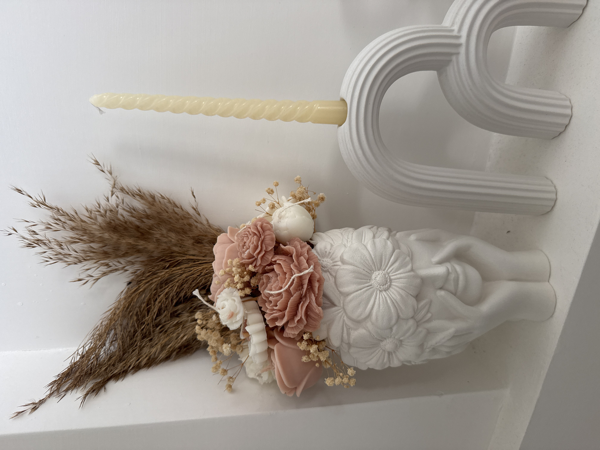 The image shows a still life arrangement on a white shelf. On the left is a white vase shaped like a woman's face with flowers adorning her head. The vase holds a bouquet of dried grasses and several pink and white floral-shaped candles. To the right of the vase is a white arch-shaped candle holder with a single twisted yellow candle inserted. The background is a plain white wall. The image does not contain any information to suggest a location in Rome, Italy.