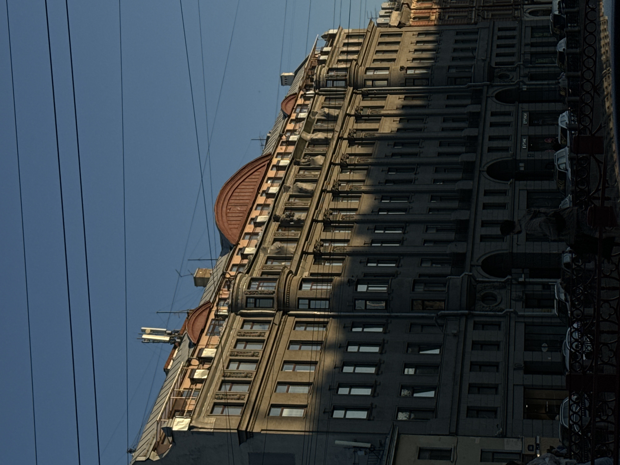 The image captures a view of a grand, historic building in Saint Petersburg, Russia, bathed in the warm, angled light of the late afternoon sun. The building is a multi-story, ornate structure with a distinct mansard roof. It features numerous windows, balconies, and architectural details, including columns and decorative carvings. The façade is rendered in a muted grey stone. On the rooftop, several white antennas are visible. Overhead, power lines crisscross the clear blue sky. In the foreground, a dark, decorative metal railing runs horizontally across the bottom of the frame. Beyond the railing, several cars are parked or moving along a street. The overall impression is one of a classic urban European setting, typical of Saint Petersburg's historical districts.