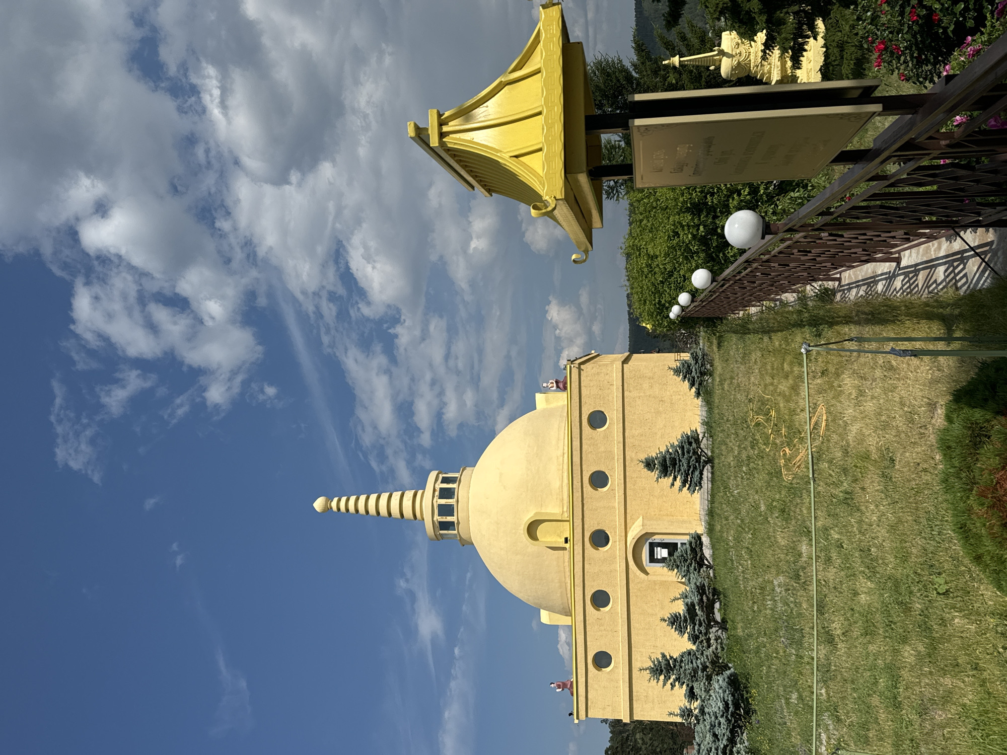 The image captures a bright, sunny day at the Ivolginsky Datsan, a prominent Buddhist monastery located near Ulan-Ude, Russia. The central subject is a large, yellow stupa, a dome-shaped religious structure, topped with a spire. The stupa has several circular windows and is adorned with small statues of figures on its upper level. In front of the stupa, a line of miniature evergreen trees is planted. To the right of the stupa, a smaller, yellow pagoda-like structure stands on a pole, with a sign beneath it. A brown metal fence with spherical lights runs along a walkway, leading the eye towards the stupa. Lush green grass covers the foreground, with some yellow and green hoses visible on the lawn. The sky above is a vibrant blue, dotted with white, fluffy clouds. In the background, trees and hills can be seen, suggesting the monastery is situated in a natural, somewhat rural setting. The overall impression is one of peace and spiritual significance, set against a beautiful natural backdrop.