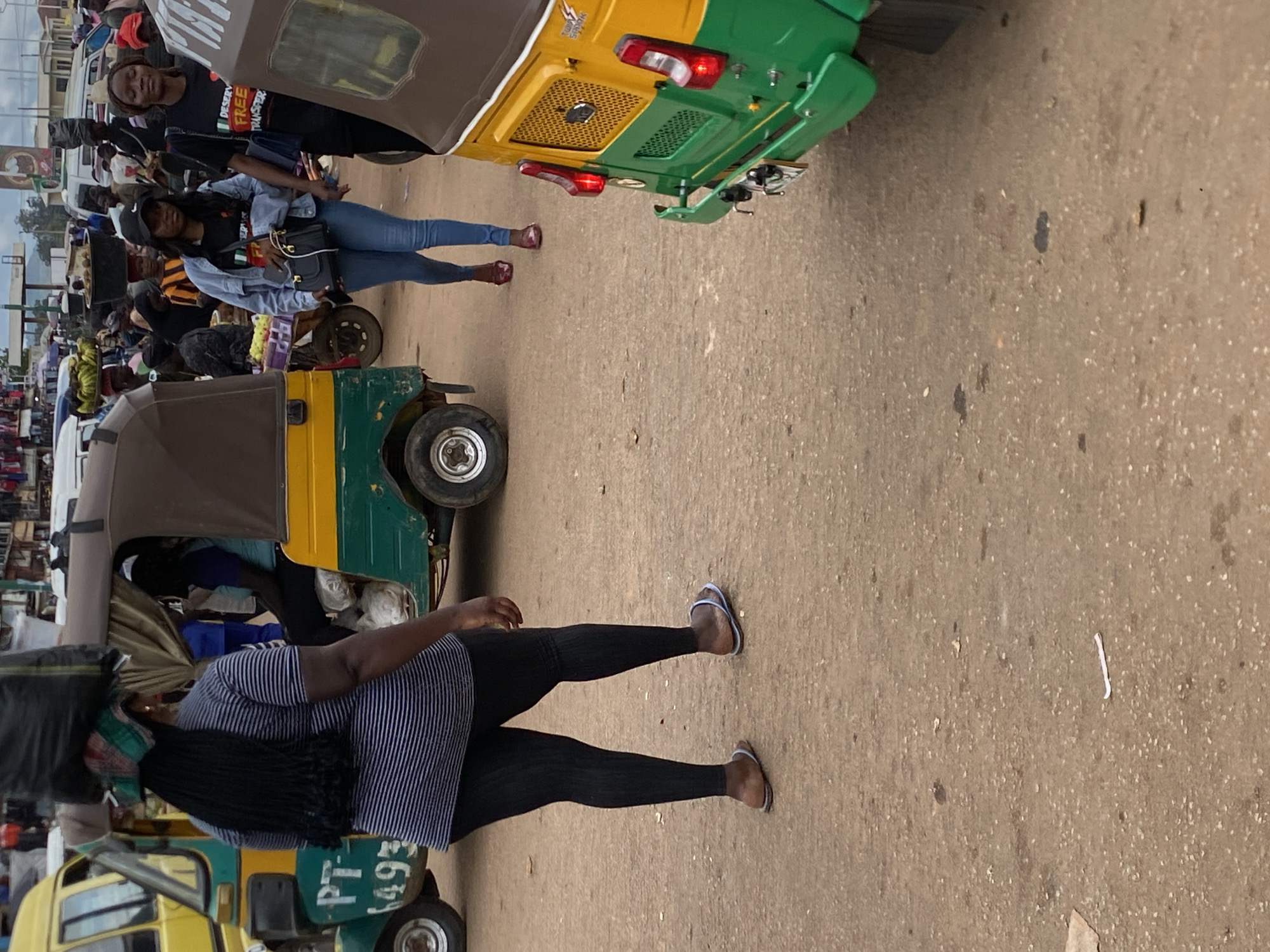 The image depicts a street scene in Bukuru, Nigeria, featuring a woman carrying a large bundle of items on her head, walking past a green and yellow auto-rickshaw. She is wearing a striped long-sleeved shirt and black leggings. To the right, another woman is walking in a denim jacket and jeans, carrying a black purse. In the foreground, the rear of a green and yellow auto-rickshaw is visible. The ground is made of dirt and gravel. The background shows a bustling market area with various vehicles and people.