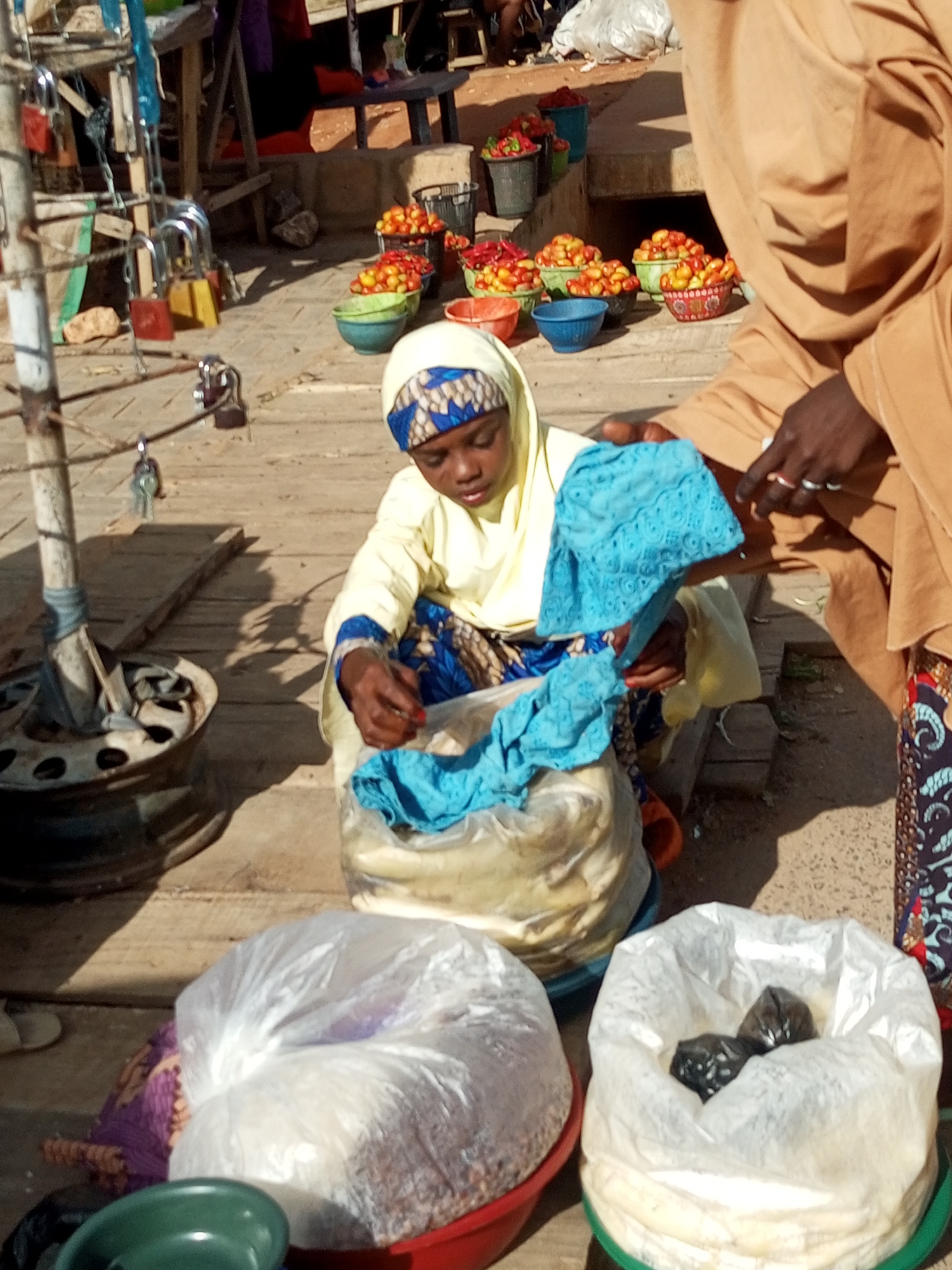 This is a street market scene in Tempure, Nigeria. A young person, likely a child, wearing a yellow garment and a patterned headscarf is kneeling and interacting with a large, clear plastic bag filled with a light-colored substance. They are holding a piece of blue, patterned fabric. Another person, partially visible and wearing a tan-colored robe and patterned trousers, is standing to the right, with their hands near the young person. In the background, there are various colorful bowls filled with small, round fruits, likely tomatoes. There are also several padlocks hanging from a metal structure on the left side of the image. The ground appears to be made of wood planks and dirt.