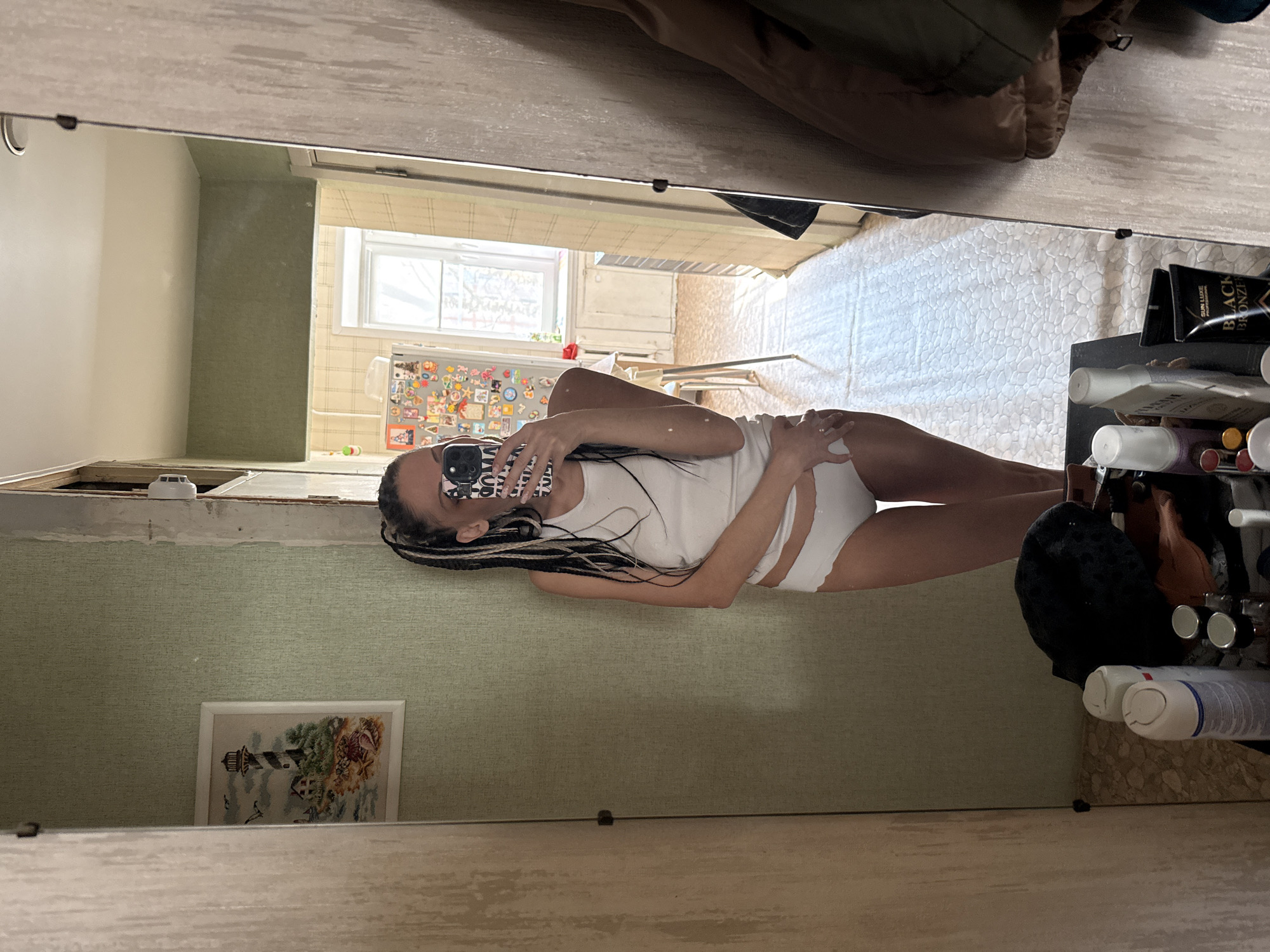 A woman is seen in a mirror, holding a smartphone in front of her face, with a white tank top and underwear. The reflection includes the interior of a room with light green walls and a picture hanging on the wall. Further into the reflection, a kitchen area can be seen with a window. In the foreground, next to the mirror, are various cosmetic products and other items on a surface. The location is likely Rusanovo, Russia.