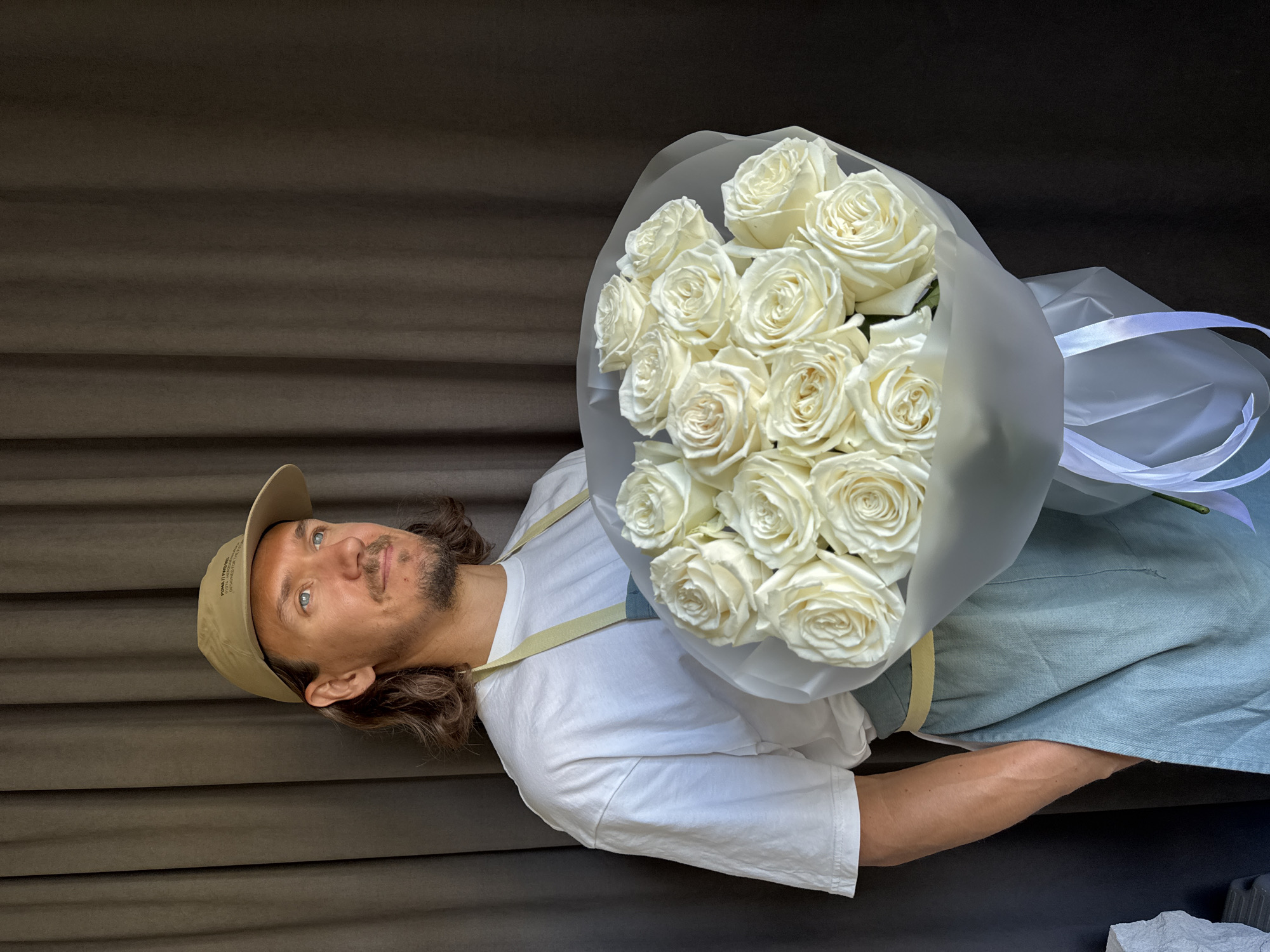 A man wearing a light brown baseball cap, a white t-shirt, and a light blue apron stands against a dark brown textured background. He is holding a large bouquet of white roses wrapped in sheer white paper and tied with white ribbon. The man has light brown, wavy hair and a goatee. He is looking slightly to his left, with a gentle smile. The bouquet of roses is the focal point, with many full blooms. The overall lighting suggests daytime. The image appears to be a studio shot or a staged photograph, with the background being a dark, textured fabric. There is text visible on the man's cap, but it is too blurry to read. The setting is likely indoors given the controlled lighting and background. No specific location cues point to Izhevsk, Russia. The mood is calm and pleasant.