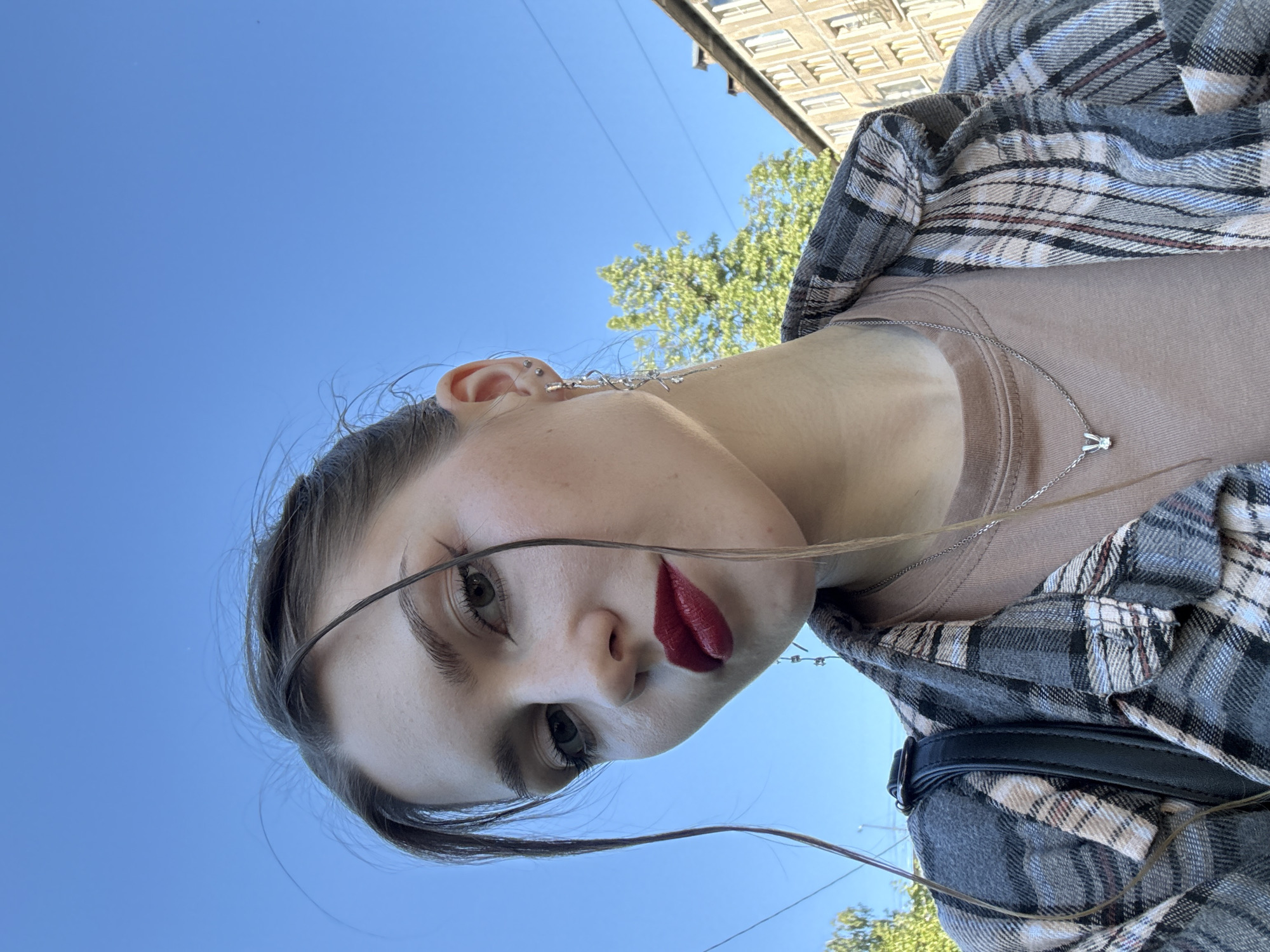 A young adult female takes a low-angle selfie outdoors in Saint Petersburg, Russia, against a clear blue sky. She looks directly at the camera with a slight tilt of her head. Her face features prominent eyebrows, mascaraed eyelashes, and dark red lipstick. Her dark brown hair is styled with loose strands framing her face. She wears a light brown crew-neck t-shirt layered under an open grey, white, and red plaid flannel shirt. A black strap, likely from a backpack, is visible over her left shoulder. On her visible right ear, she has a helix piercing with a small stud and two lobe piercings; one with a small stud and the other with a long, silver, twisted dangling earring adorned with small jewels. A thin silver chain necklace with a small bunny pendant is visible around her neck. The background features bright blue sky, with green deciduous trees visible on both sides. On the far right, a light-colored, multi-story building with multiple rectangular windows, consistent with older European urban architecture, is partially visible. Thin power lines traverse the upper right portion of the sky.