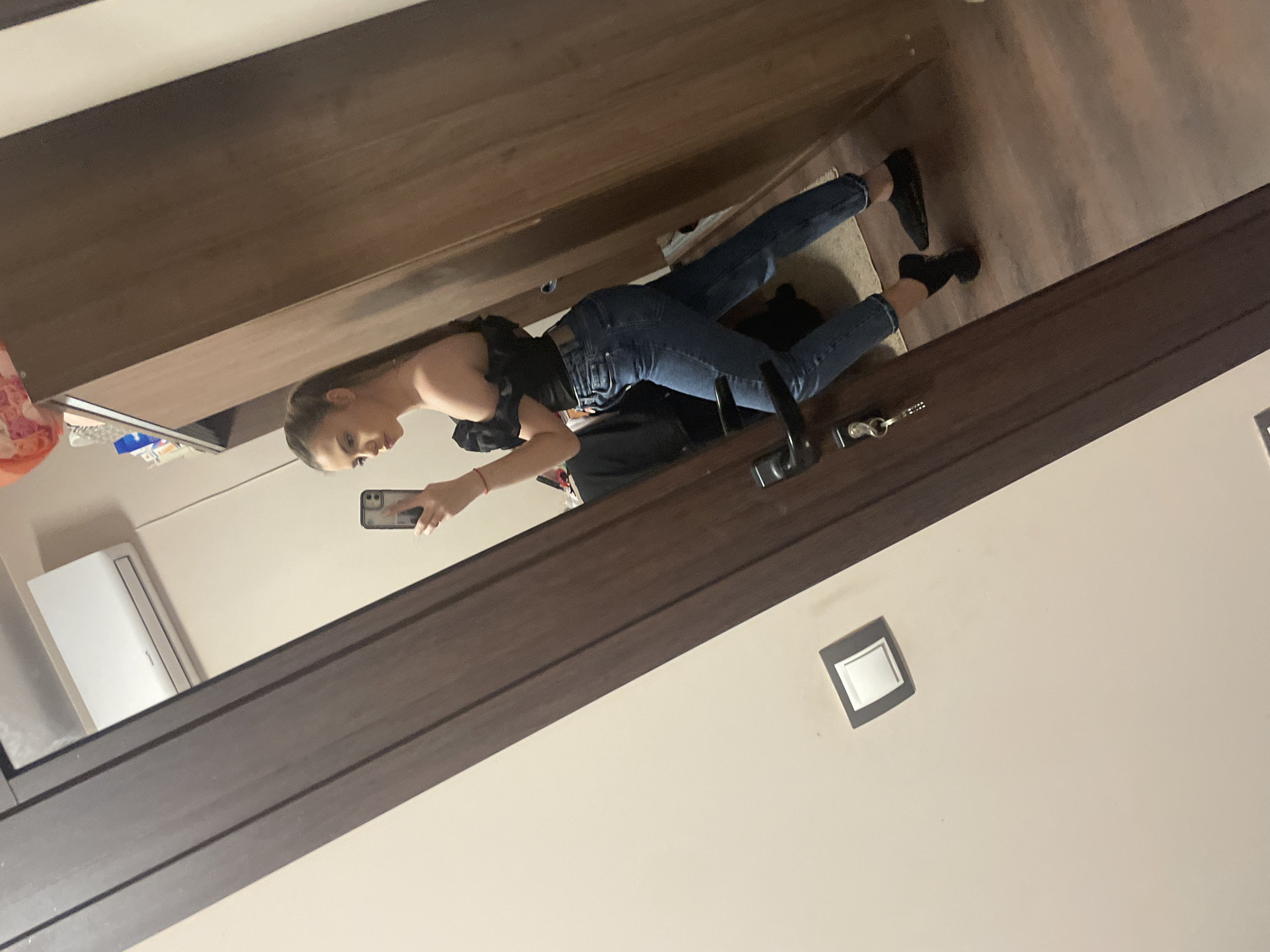The image shows a woman taking a selfie in a mirror. She is wearing a black top and blue jeans. She is standing in a room with wood paneling and a light switch on the wall. There is also an air conditioner unit visible in the reflection. The location appears to be inside a building in Unknown, Unknown.