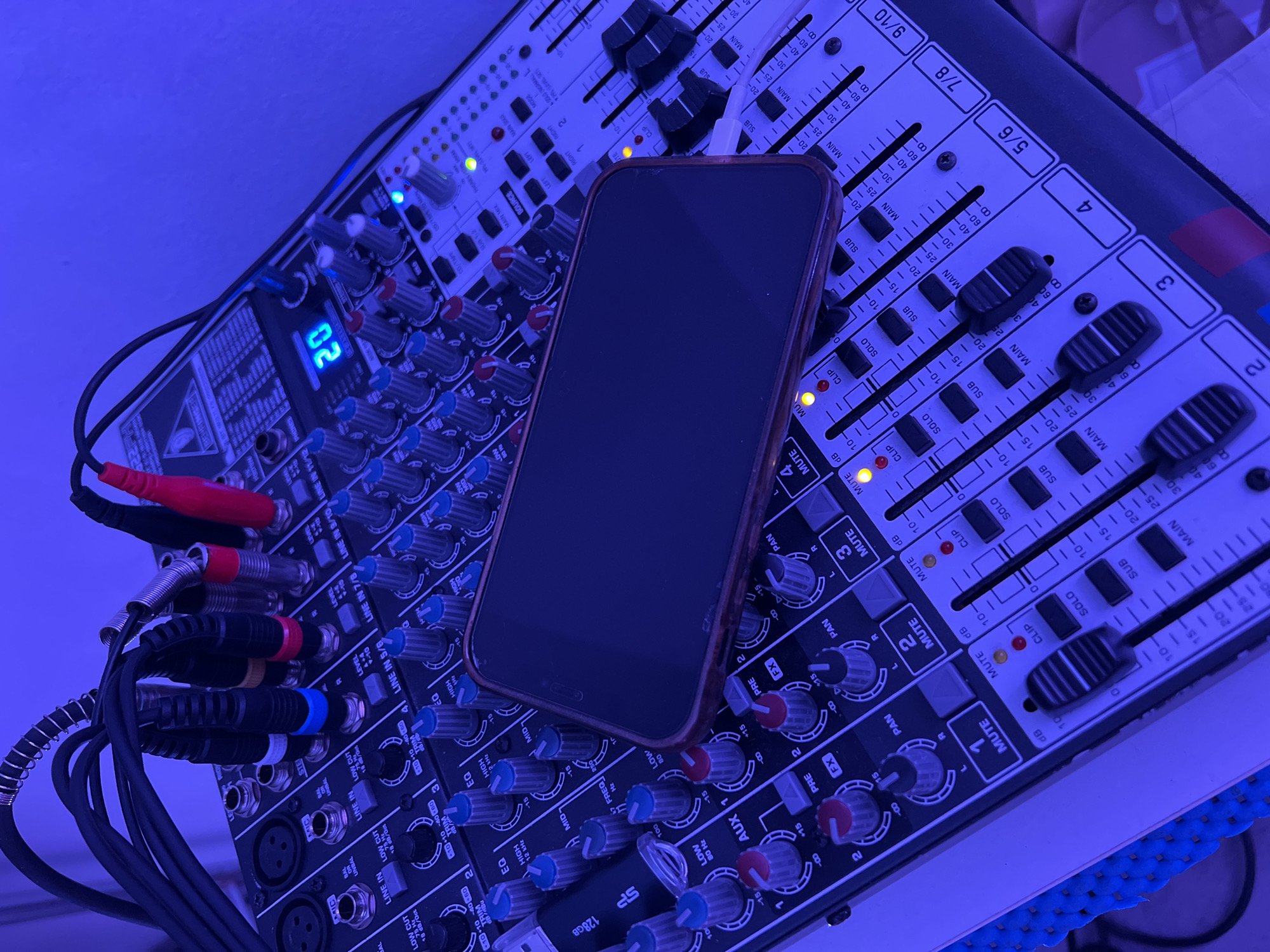 In Santeramo in Colle, Italy, a Behringer audio mixing console is captured in an overhead, slightly angled view, illuminated by strong blue/purple ambient lighting. Numerous audio cables, including XLR and 1/4-inch jacks with black, blue, yellow, and red connectors, are securely plugged into the upper rear input panel of the console. A smartphone, equipped with a red/brown protective case, rests face down on the central section of the mixer, positioned over several control knobs. To the left, a "SP" branded 128GB USB flash drive lies on the console's surface.
The mixer is powered on, indicated by an illuminated digital display showing the number "20" and various active LED indicators, including red "CLIP" lights, yellow "MUTE" lights, green level meters, and blue "SOLO" lights. The console features multiple input channels, each with a series of blue, red, and grey rotary knobs controlling parameters such as EQ (High, Mid, Low), Trim, Aux, FX, and Pan. Black fader controls are present for individual channel levels, labeled from 1 to 9/10, alongside corresponding "MUTE" and "SOLO" buttons. A blue textured mat is visible beneath the left edge of the console. No individuals are present in the scene.