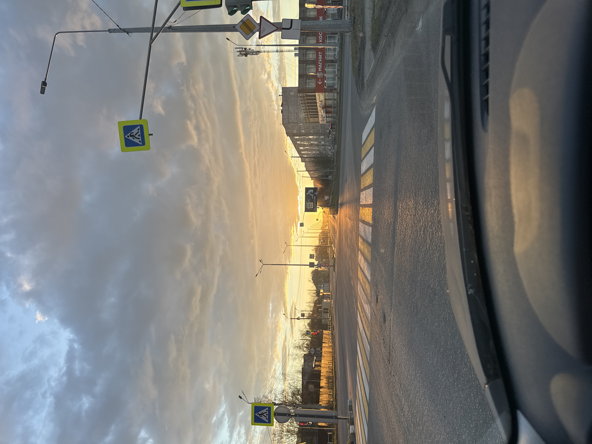 The image depicts a street in Apatity, Russia, during a sunset. The perspective is from inside a vehicle, looking out onto the street. The road has a crosswalk marked with white and yellow stripes. Streetlights line the road on both sides. Traffic signals are visible, as well as pedestrian crossing signs. Buildings are on the right side of the street. The sky is filled with clouds and illuminated by the setting sun. The scene is static; no people or moving vehicles are observed.