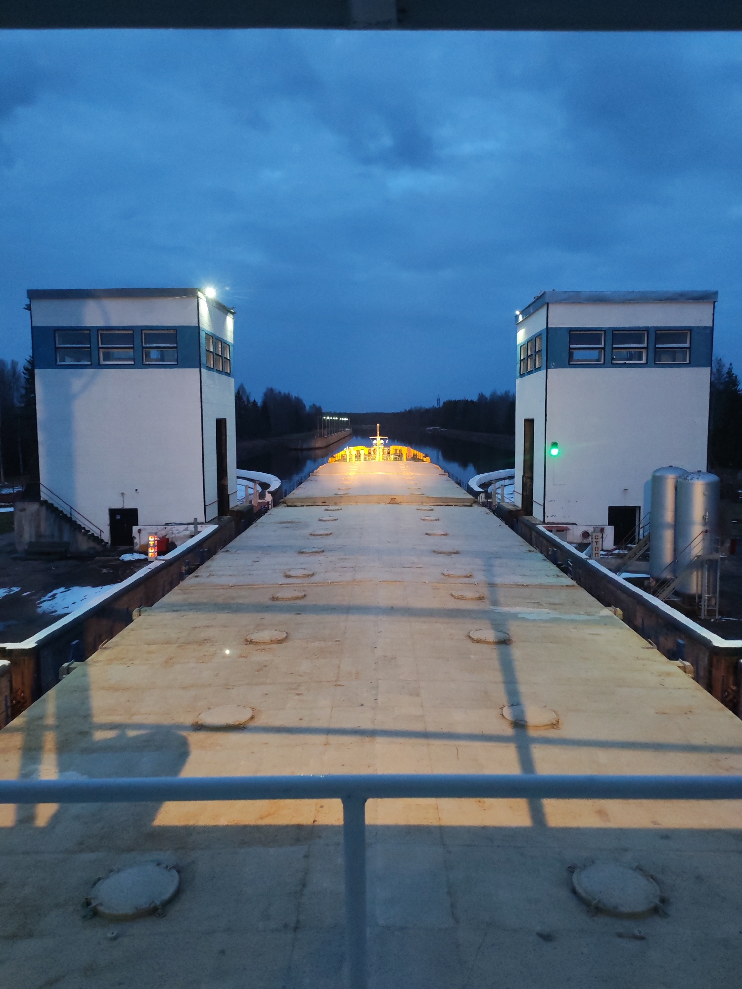 A high-angle view shows a canal lock system at dusk, possibly in the city of Unknown, Unknown. The lock structure features two identical, white, two-story buildings with blue accents at each end of the platform. Each building has a set of windows on its upper level. A light shines atop each building. A wide, concrete platform stretches between the buildings, marked with circular access points. The platform appears to be the top of a lock. Beyond the lock, a narrow canal extends into the distance, lined with trees. A barge or boat with lights can be seen in the canal beyond the lock. The sky is a deep blue with scattered clouds. A metal railing is visible in the foreground, suggesting the photograph was taken from an elevated position. Patches of snow remain on the ground near the buildings. A green light is visible on the right building. Cylindrical tanks are present to the right of the right building. A set of stairs leads down from both of the buildings.