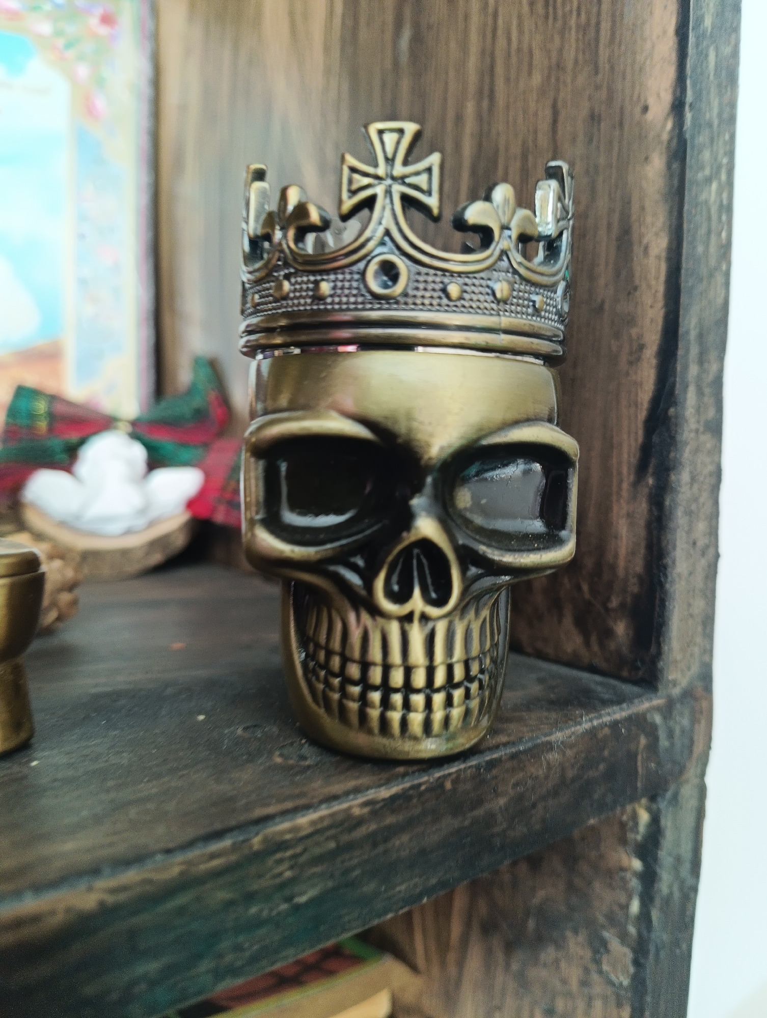 The image depicts a close-up view of a metallic skull-shaped object with a crown on its head, placed on a dark wooden shelf. The object is golden in color and features intricate detailing, particularly on the crown and the skull's teeth and eye sockets. In the blurred background, a white angel figurine is visible, adorned with a red and green plaid ribbon. The shelf itself is part of a wooden structure with a weathered appearance. The setting appears to be indoors, possibly a decorative display or collection. The location in Karnobat, Bulgaria is not discernible from the visual information provided in the image.