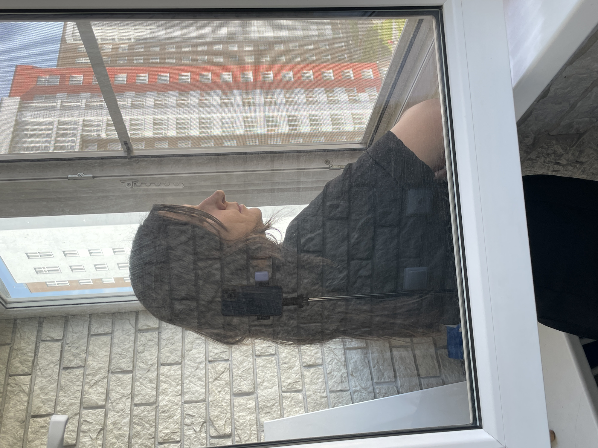 A medium shot of a woman's reflection in a window. The woman has long, dark hair and is wearing a dark gray shirt. She is facing to the left. A cell phone and cable are reflected on her head. The background shows a brick wall in the lower left corner and a large building with many windows in the upper portion of the image. The building has a red stripe on the top. The window frame is white. The location is Unknown, Unknown.