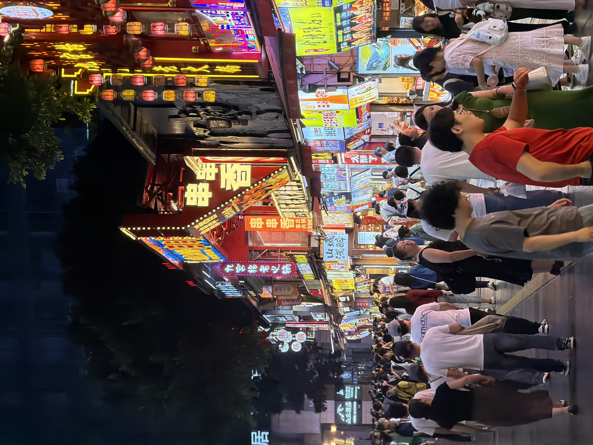 The image depicts a bustling night market scene in Yuzhong District, China. A dense crowd of people is walking along a paved street lined with brightly lit stalls and shops. The architecture features traditional Chinese elements with glowing neon signs and red lanterns adorning the buildings. The stalls display a variety of goods, likely food and merchandise, with colorful banners and signs in Chinese characters. The lighting is a mix of the warm glow from lanterns and the harsh, vibrant colors of neon advertisements. The people are dressed in casual attire, suggesting a warm evening. Some individuals are looking at the stalls, others are conversing, and many are simply moving through the crowded thoroughfare. The overall atmosphere is lively and energetic, indicative of a popular urban night market.