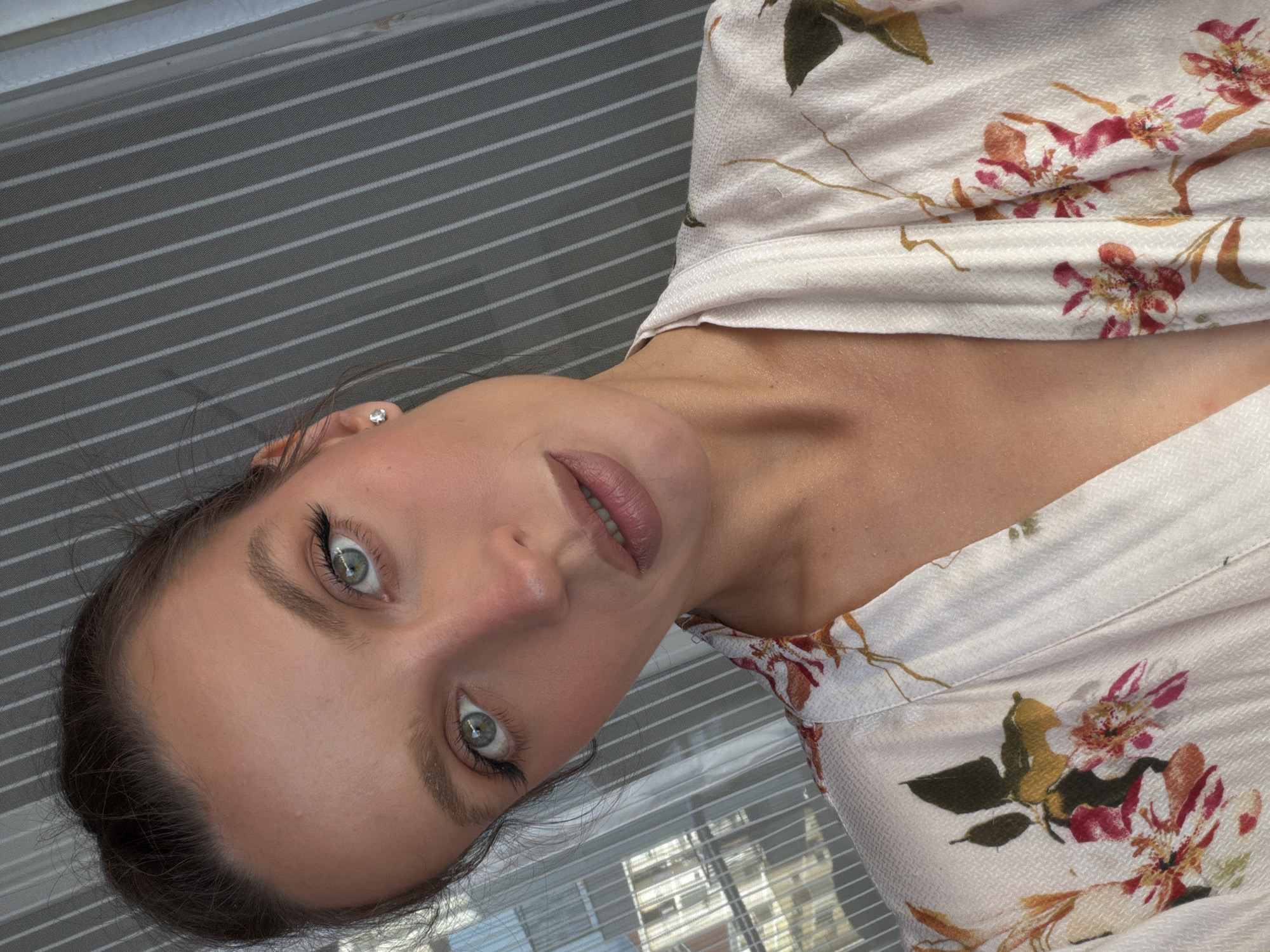 A young woman with fair skin, light green eyes, and dark hair is looking directly at the camera. She is wearing a white shirt with a floral pattern. An earring is visible in her left ear. The background is a blurry view of a window with horizontal blinds. There is no definitive evidence in the image to suggest the location is in Budva, Montenegro.