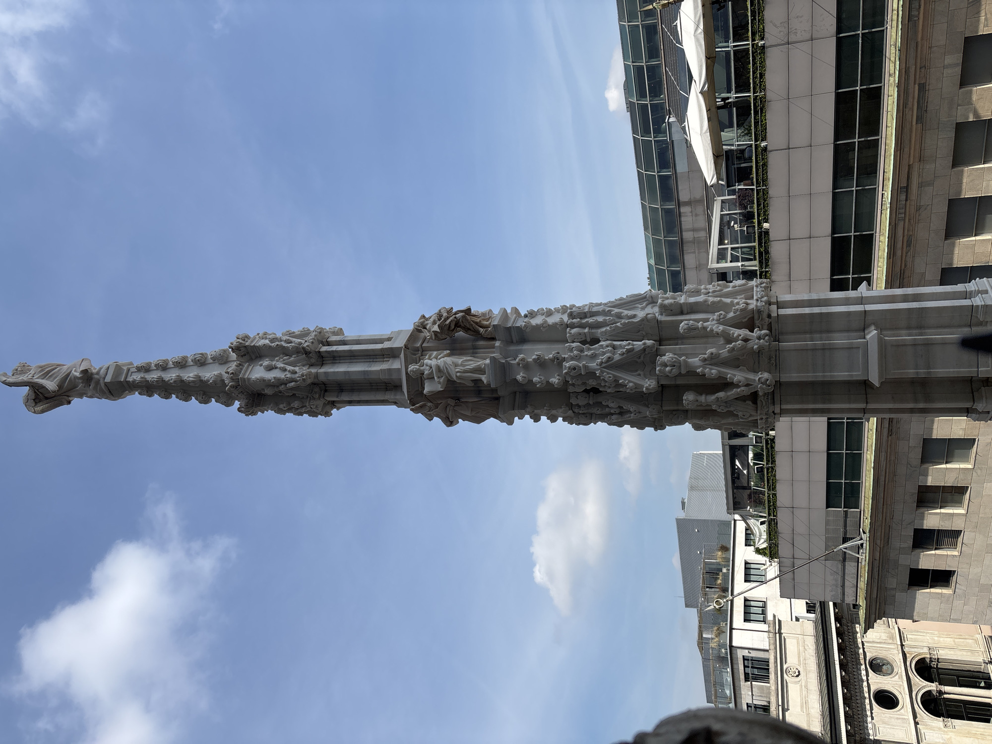 The image shows a detail of the Milan Cathedral (Duomo di Milano) in Milan, Italy. Specifically, it depicts the exterior spires and sculptures of the cathedral against a backdrop of a bright blue sky with light clouds and adjacent urban buildings. The architectural details of the spire are prominently displayed. Modern buildings can be seen adjacent to the cathedral, suggesting the juxtaposition of historical architecture with contemporary urban elements. The foreground and middle ground is occupied by the marble spire, while the background shows modern buildings. There are no people visible in this shot.