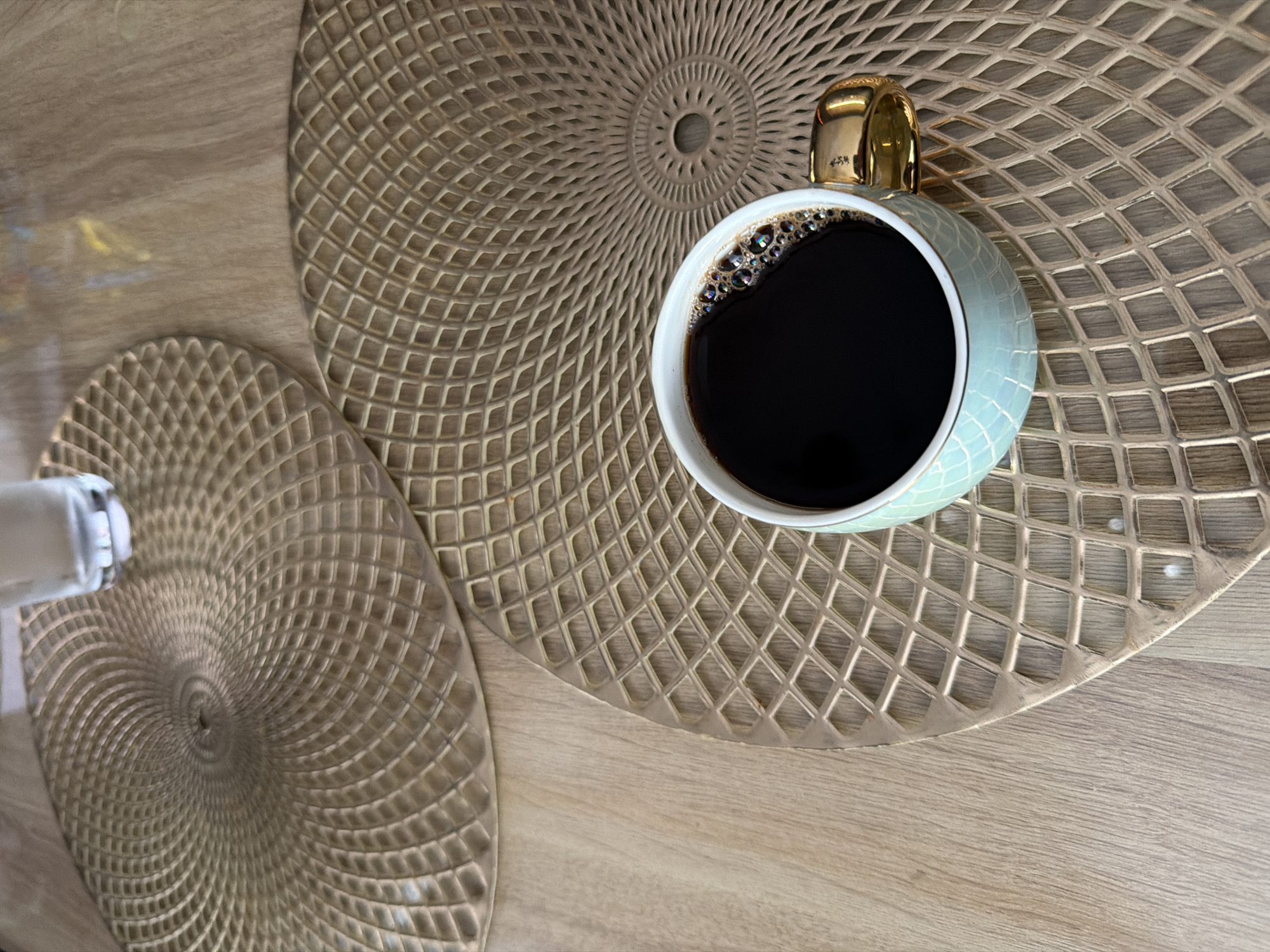 A close-up shot captures a cup of dark coffee with frothy bubbles resting on a decorative placemat. The placemat, made of a woven material with a diamond pattern, is gold or brass colored and has a circular, radiating design. Another similar placemat is partially visible in the upper left of the frame, and beyond that, a glass of water. The surface beneath the placemats appears to be a light-colored wooden table. The coffee cup itself is light blue with a textured surface and a shiny gold handle. The scene is likely indoors, possibly in a dining or living area.