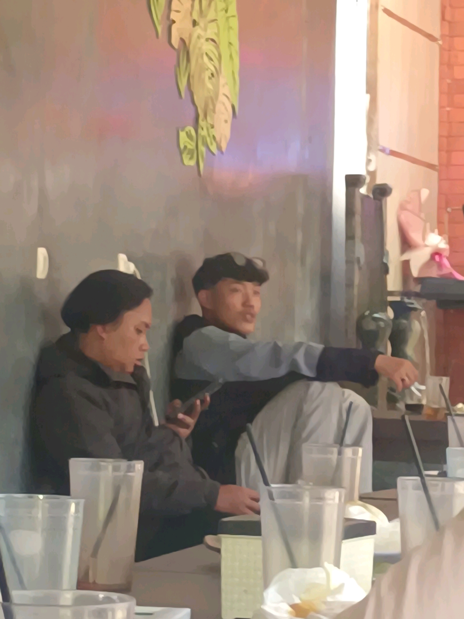In Kerangkulon, Indonesia, two individuals are seated in what appears to be an indoor dining establishment. A person with dark hair and wearing a dark jacket is looking down at a mobile phone held in their left hand. Next to them, a person wearing a dark cap and a blue and grey jacket is looking to their right and appears to be speaking, with their right arm extended. In the foreground, several clear plastic cups and a white napkin are visible on a table. The background features a textured wall with decorative plant-like cutouts and a brick wall. The overall lighting suggests an indoor setting, possibly during the evening or in a dimly lit area.