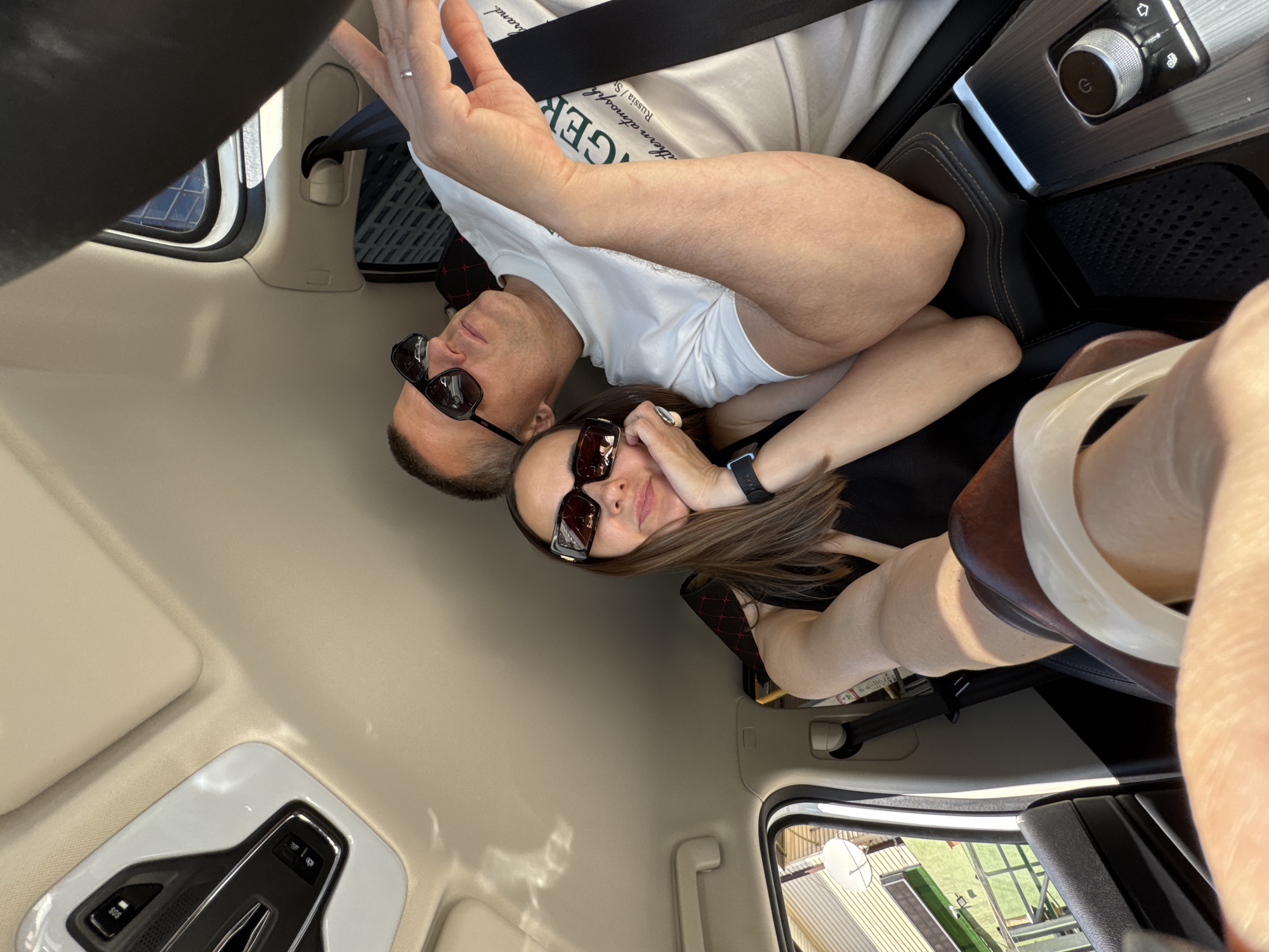 The image depicts the interior of a vehicle, possibly in the city of Sochi, Russia, with two individuals occupying the front seats. The photograph is taken from a low angle, likely from the passenger side, looking forward and towards the center of the car.
On the left, a female subject is seated in the front passenger seat. She has long brown hair and is wearing dark, rectangular sunglasses and a black sleeveless top. A dark digital watch is visible on her left wrist. She rests her chin on her right hand, which is positioned on her lap or an armrest, while her left arm is bent. A light-colored, possibly translucent, bracelet or ring is visible on her right hand in the foreground. Through the open window on the left, a beige-colored building with horizontal siding and a white satellite dish are discernible under sunlight.
On the right, a male subject is seated in the driver's seat. He has short hair and is wearing dark, rectangular sunglasses and a white short-sleeved t-shirt with dark green text, including partial words like "GERGERS" and "southern atmosphere. Russia / S...". He is secured by a black seatbelt across his chest. His head is tilted slightly back, and he is looking upwards and to the right. His left arm is bent at the elbow, with his hand raised and fingers slightly splayed, appearing to point or gesture towards the upper right of the frame. A silver-colored ring, consistent with a wedding band, is present on his left ring finger. Through the window on the right, reflections and partial views of modern urban buildings with dark glass facades are visible.
The car's interior features a beige headliner and sun visors. An overhead console unit, white with black accents, is visible on the ceiling, displaying buttons including one labeled "SOS" and a lock icon. The center console between the two individuals is dark-colored, featuring a metallic-trimmed rotary dial with a power symbol (likely for ignition or gear selection) and adjacent control buttons. A textured, possibly rubberized, section is also visible on the console. The car seats are dark, and a hint of red stitching is visible on the headrest behind the male driver.