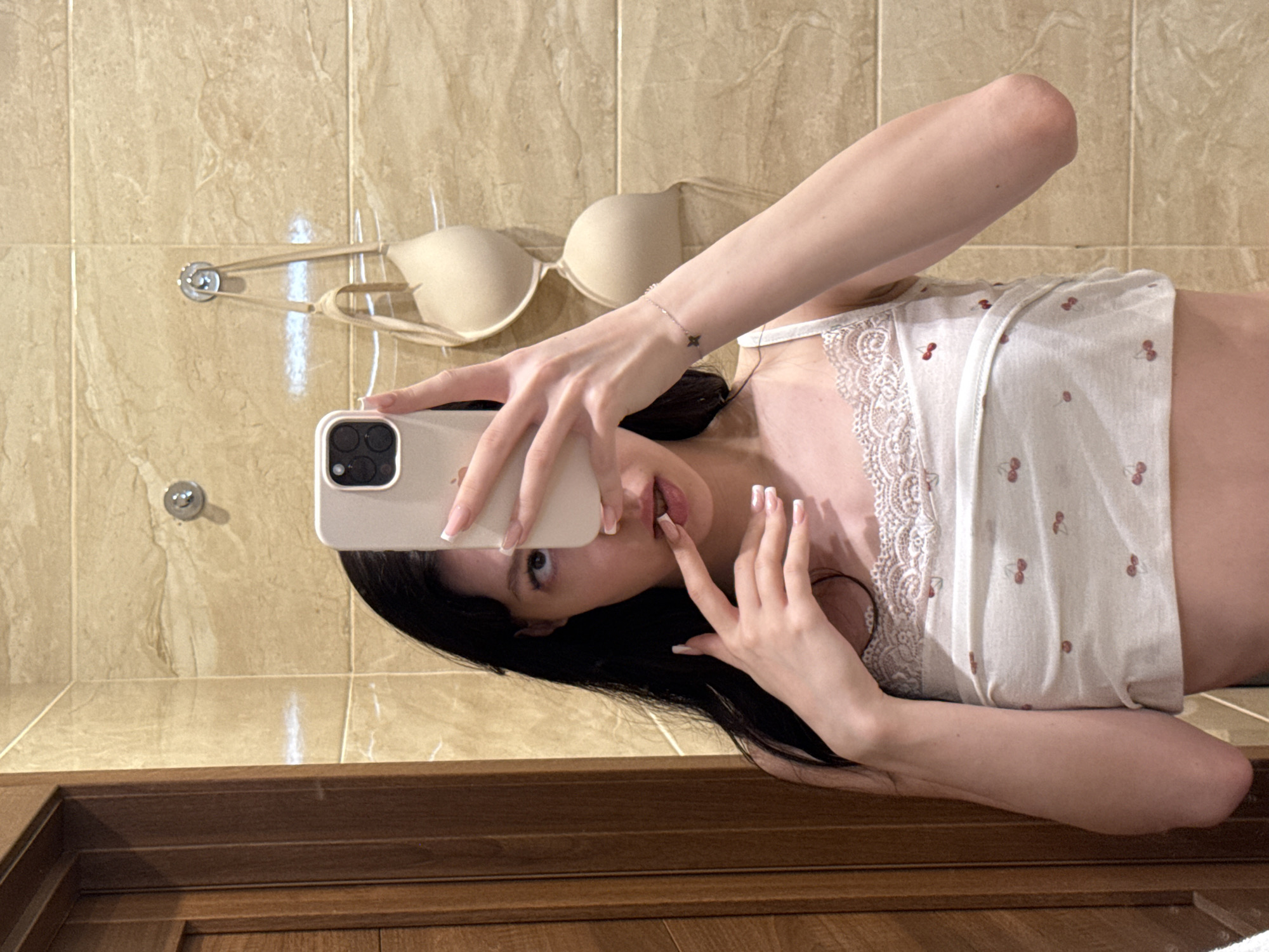 A young woman with long, dark hair takes a selfie in a bathroom. She is wearing a white camisole with a cherry pattern and lace trim. Her right hand is holding a smartphone, obscuring part of her face, while her left hand is raised to her lips. A beige bra is hanging on a hook on the tiled wall behind her. The room appears to be a domestic bathroom, likely in an apartment in Khimki, Russia.