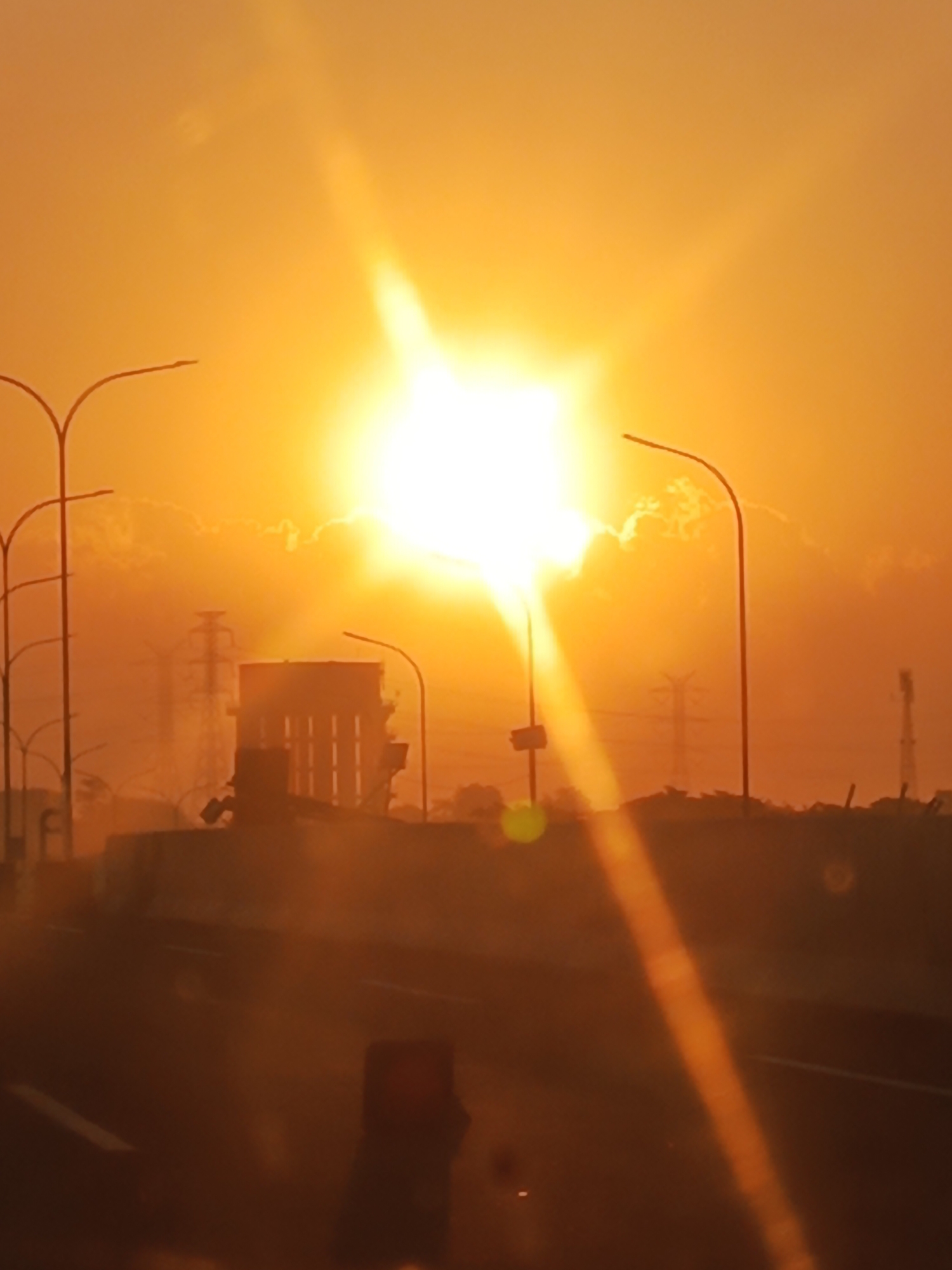 An outdoor, medium shot captures a sunset over a highway in Gempol, Indonesia. The sun, a bright, luminous orb, dominates the sky, casting a warm, orange glow over the entire scene. Lens flare streaks across the image, originating from the sun and extending downwards. In the background, a large, rectangular structure with vertical lines is visible, possibly a building or industrial facility. Several streetlights with curved poles are scattered throughout the frame, silhouetted against the bright sky. Power lines and towers are also discernible in the distance, indicating an urban or industrial environment. The foreground shows a highway with lane markings, and a dark, out-of-focus object, possibly a vehicle or a person, is present in the lower portion of the image. The overall atmosphere is hazy, with a subtle glow emanating from the sun, suggesting the presence of dust or moisture in the air.