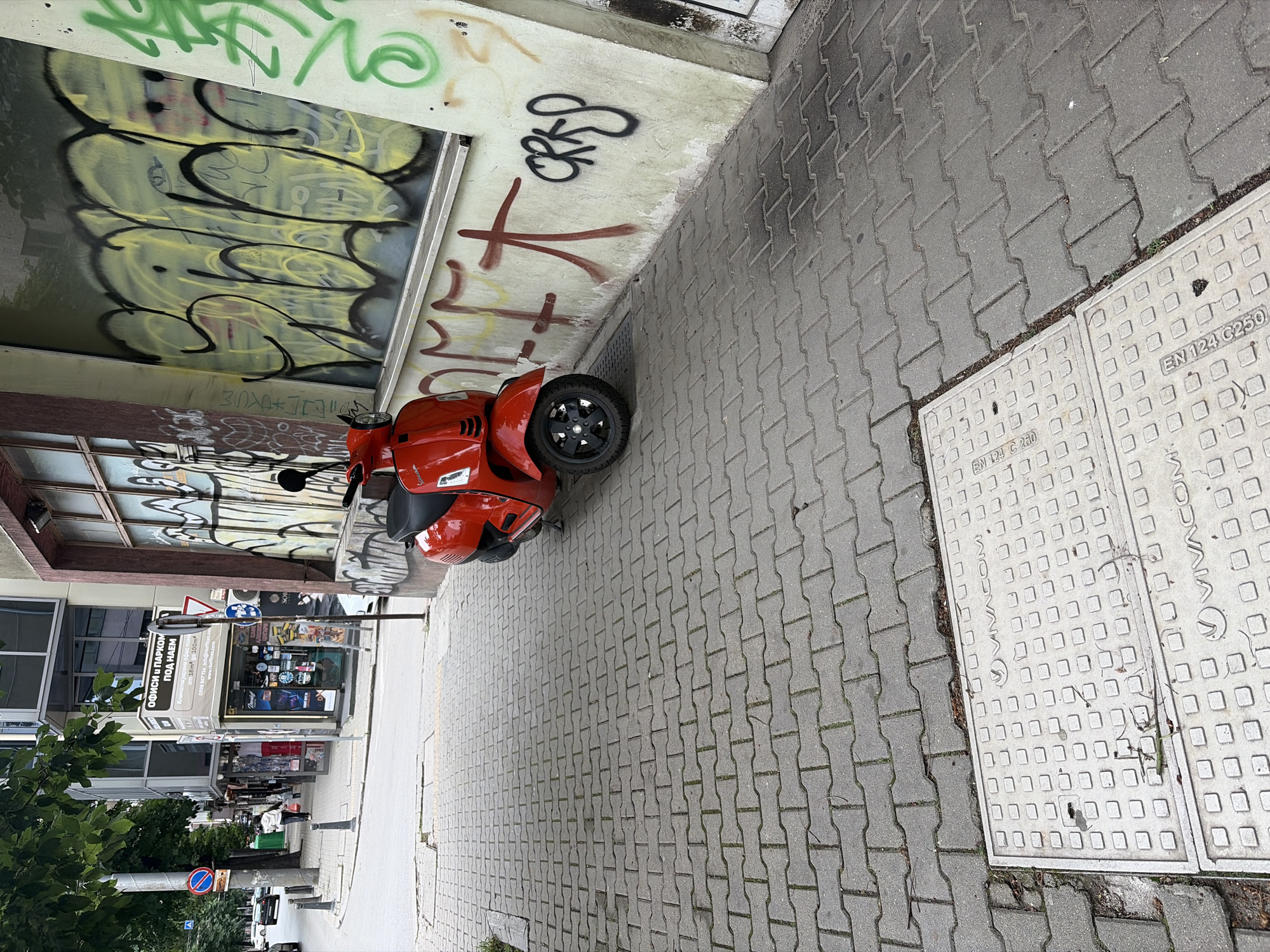 The image captures a street scene in Sofia, Bulgaria, with a focus on a red Vespa scooter parked on a grey brick sidewalk. The scooter is positioned in front of a building adorned with extensive graffiti. The graffiti features large, yellow stylized lettering with black outlines and some red markings. To the left of the scooter and building, a sidewalk runs alongside a street, with trees and other buildings visible in the background. A manhole cover, marked "VIVACOM" and "EN 124 C 250", is prominent on the sidewalk in the foreground. The overall impression is an urban environment with elements of commercial activity and artistic expression through graffiti.