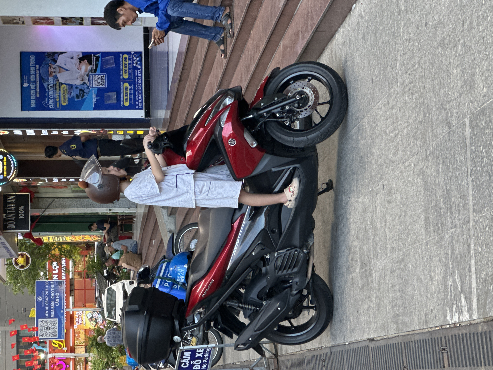 The image shows a person wearing a helmet and white clothing sitting on a red and black motorcycle parked on a sidewalk in Bac Nha Trang Ward, Vietnam. The sidewalk is adjacent to a building with various businesses. In the background, other people are visible, along with street signs, restaurants, and a dental clinic poster. A man is standing on stairs looking at his cell phone. The image is rotated.