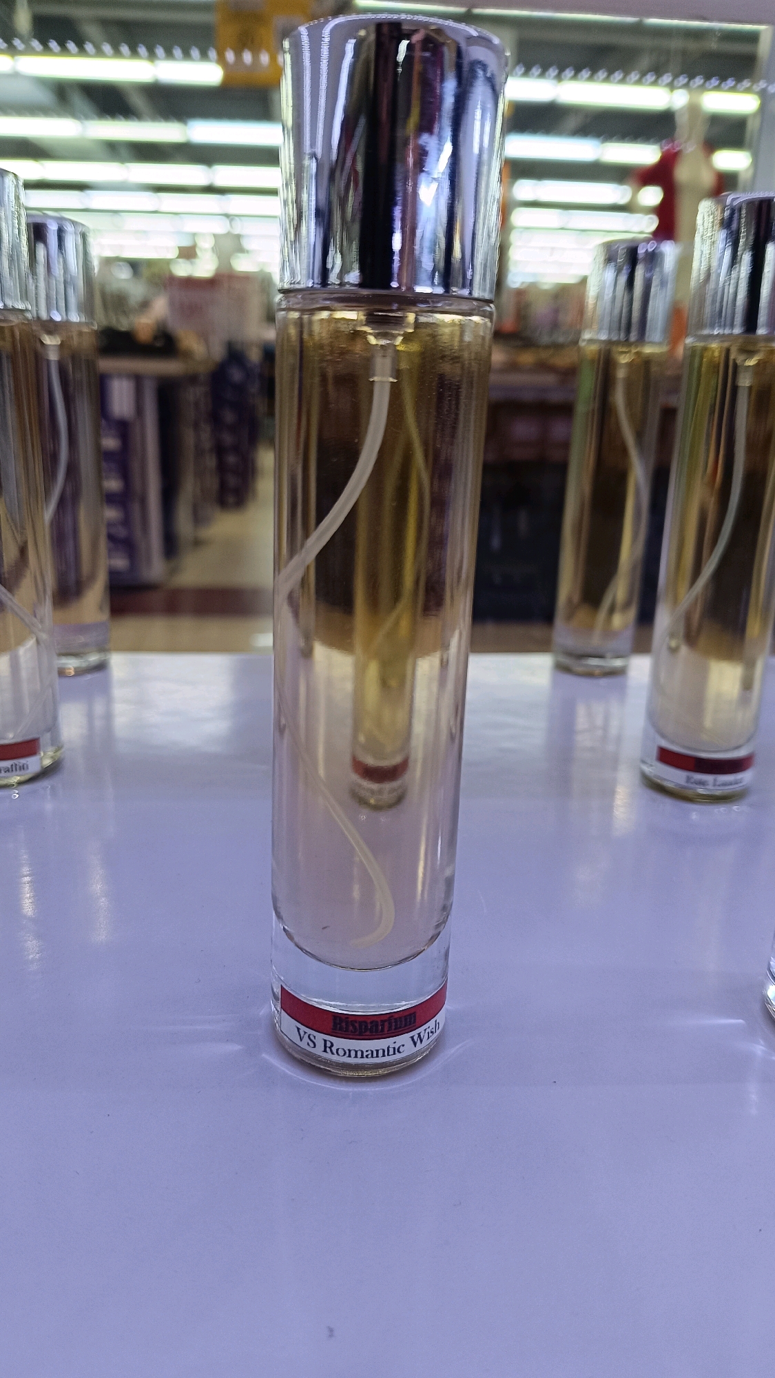 The image shows a close-up, eye-level view of several perfume bottles on a white surface. The bottles are tall and cylindrical, with clear glass and silver caps. The perfume inside is a light golden color. A white tube extends from the cap into the liquid. One bottle in the foreground has a red label with white text that reads "Risparfum VS Romantic Wish." In the background, shelves with various items are visible, suggesting a retail environment, possibly a store or market in Tomohon, Indonesia. The lighting is bright, with fluorescent lights overhead. There are no people clearly visible in the image.
