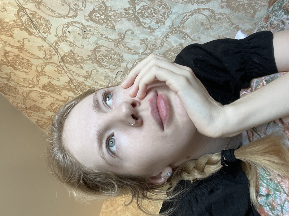 A fair-skinned person with blonde hair in a braid is looking up. Their hand is near their face, resting on their cheek. They have a nose ring and an earring. The person is wearing a black, short-sleeved shirt with a floral pattern underneath. The background consists of patterned wallpaper. The image appears to be taken from a slightly elevated angle.