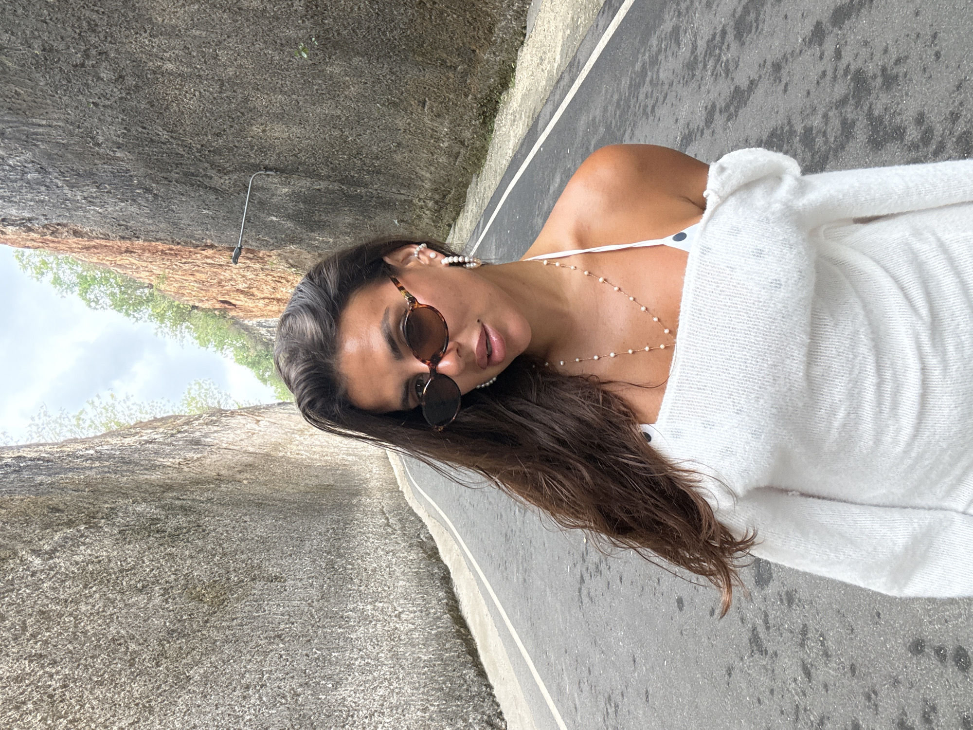 A medium shot captures a woman standing on a paved road between two imposing, tall rock walls. The road has a white line marking its center and curves to the left in the background. The rock walls, textured and uneven, rise steeply on either side of the road, with some vegetation visible at their upper edges. The woman, facing the camera, wears sunglasses with tortoiseshell frames, a white off-the-shoulder sweater, and a delicate pearl necklace. Her long, dark hair is loose and falls around her shoulders. The lighting suggests it is daytime, with some clouds in the sky. The scene appears to be an open-air location, likely a natural or man-made cut through rock. The presence of a street lamp on the right rock wall indicates a developed area. The image was taken at the Melasti Cliff, a popular tourist spot in Kuta Selatan, Indonesia.