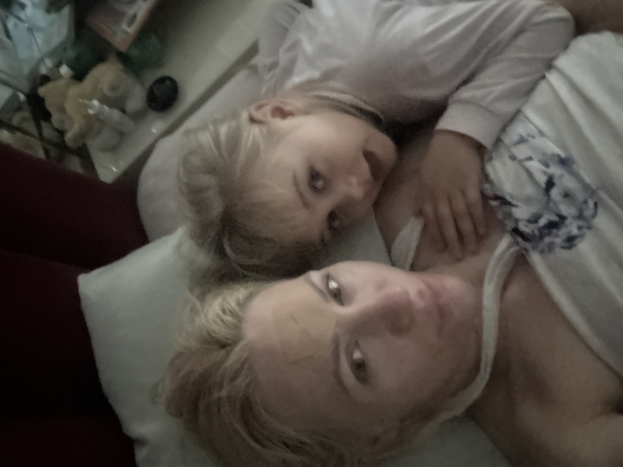 The image shows a close-up, overhead shot of two individuals, an adult and a child, lying down together. The adult, in the foreground, has light-colored hair and is looking upwards towards the camera with their eyes open. Their skin appears to have a slightly textured appearance, possibly from a facial mask. They are wearing a white tank top. The child, positioned behind the adult and slightly to the left, also has light-colored hair and is looking directly at the camera with a slightly open mouth, appearing to be speaking or laughing. The child is wearing a white long-sleeved shirt.
They are lying on a bed with white bedding, which has a subtle blue floral pattern visible on the right side of the frame. To the far left of the image, a dark reddish-brown surface, likely a piece of furniture like a couch or headboard, is visible. In the blurred background, several objects are discernible, including what appears to be stuffed animals and other household items, suggesting an indoor, domestic setting.
The lighting appears dim, implying it could be nighttime or a room with low ambient light. The overall mood is intimate and relaxed, suggesting a moment of bonding or rest between the two individuals. No visible text is present in the image.