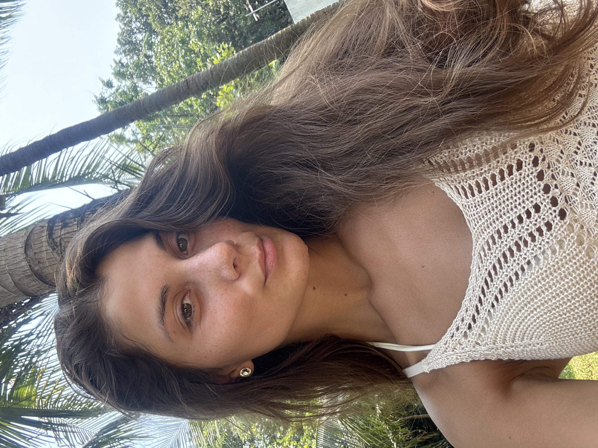 A medium shot depicts a young woman with long brown hair, positioned against a backdrop of palm trees, likely in Đà Nẵng, Vietnam. She wears a light-colored, possibly knitted, top. Her expression is neutral, and her gaze is directed towards the camera. She wears a small earring. The background showcases typical tropical foliage. The photograph is taken from a slightly low angle.
