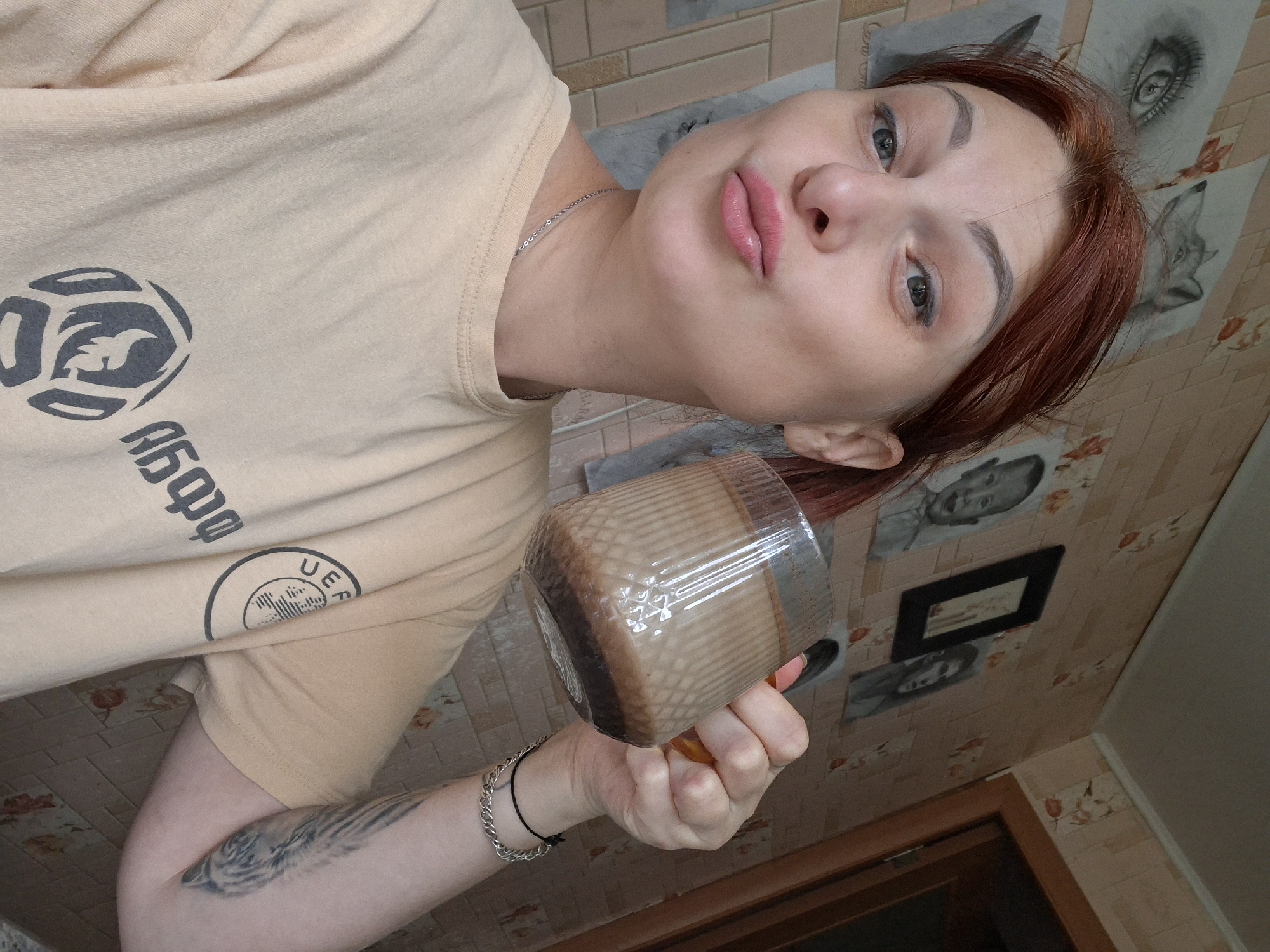 A woman with red hair is holding a mug of coffee in her right hand and making a kissy face at the camera. She is wearing a light tan t-shirt with a logo on the left side of her chest. The background appears to be a wall with a floral wallpaper pattern. There are several sketches on the wall behind her, including a drawing of an eye and a wolf. The scene is likely indoors. The specific location in the city of Myadzyel, Belarus cannot be determined from the image alone.
