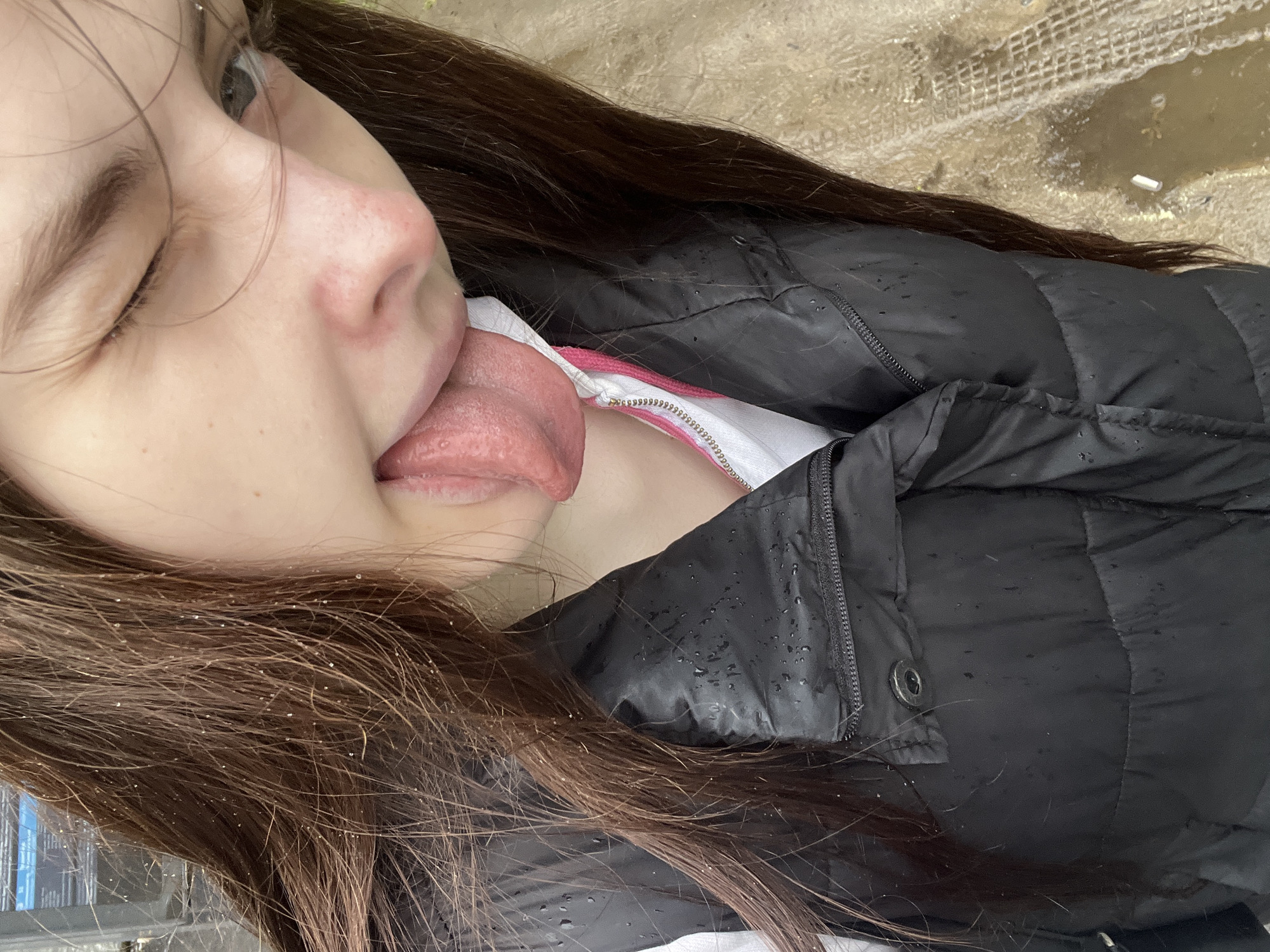 A young woman with brown hair is outdoors, wearing a black puffer jacket over a white shirt with a pink stripe, and sticking out her tongue. Her right eye is squinted shut. Her hair and jacket appear to be wet. The location is the city of Saint Petersburg, Russia.