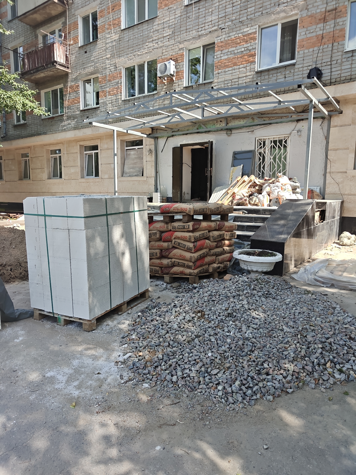 This image depicts the exterior of a multi-story brick building in Petropavl, Kazakhstan, likely undergoing construction or renovation. In the foreground, a large stack of white building blocks is neatly arranged on a wooden pallet, secured by green strapping. Adjacent to the blocks, a substantial pile of grey gravel occupies the ground. Behind the gravel, several sacks of cement are stacked on pallets, with some bearing the label "AKATER 500". A white flower pot sits near the entrance. The building itself has a facade of grey and orange brickwork, with numerous windows, some of which appear to be reflective. A metal canopy extends over the entrance, which leads to a set of stairs. A ramp with a dark, possibly granite, finish is also visible. The scene is illuminated by daylight, casting shadows on the ground, suggesting it is a sunny day. Trees and foliage are visible on the left side of the building, indicating a residential or urban environment. There are no people clearly visible in the image.