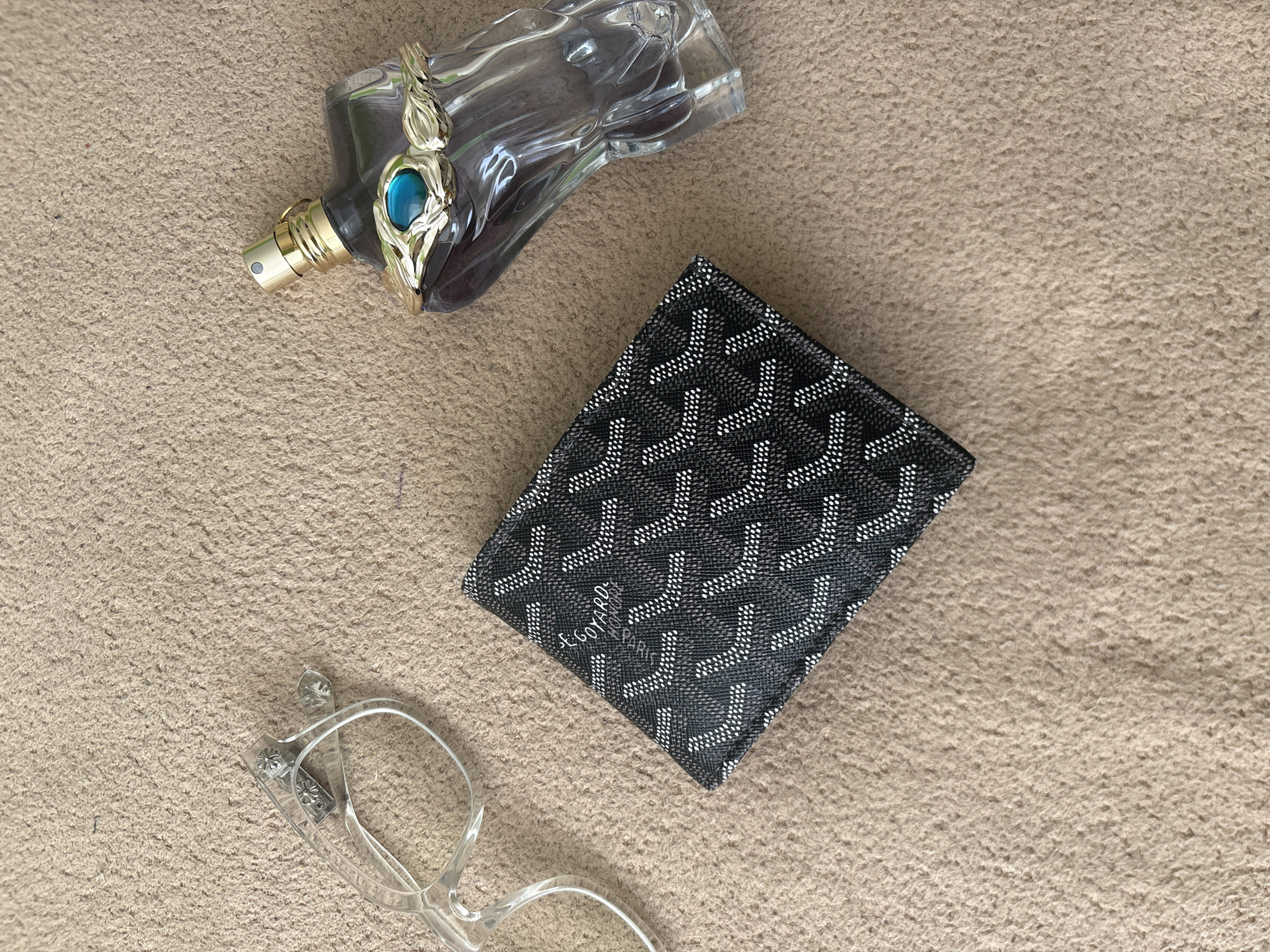 The image shows a close-up of a textured beige surface upon which several items are placed. A partially filled clear glass perfume bottle with gold accents and a blue stone is positioned at the top left. A black Goyard wallet with a repeating pattern is centered. A pair of clear plastic eyeglasses is located at the bottom left. The scene depicts personal accessories arranged on the surface. There is no information to determine the city of Stara Zagora, Bulgaria.