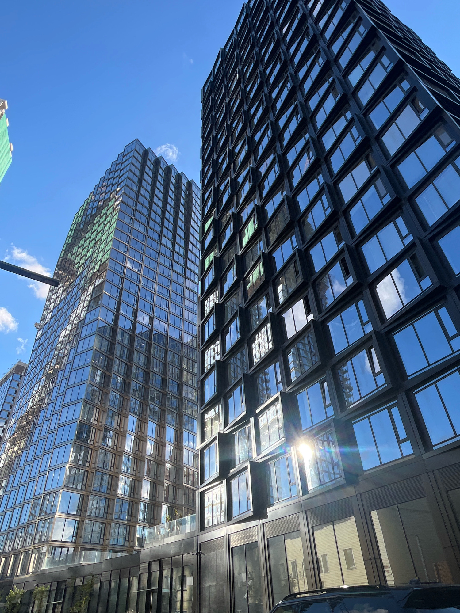 The image shows a low-angle view of modern skyscrapers against a bright blue sky with a few scattered clouds. The buildings are tall and made primarily of glass, reflecting the sky and surrounding structures. One building on the left has a slightly greenish tint due to some construction covering on its upper floors. The building on the right is darker, with a more angular design. Sunlight glints off the glass facade of the right-hand building, creating lens flare. In the foreground, a black SUV is partially visible. The scene is urban, with the architectural style suggesting a contemporary city environment. The location is identified as Moscow, Russia.