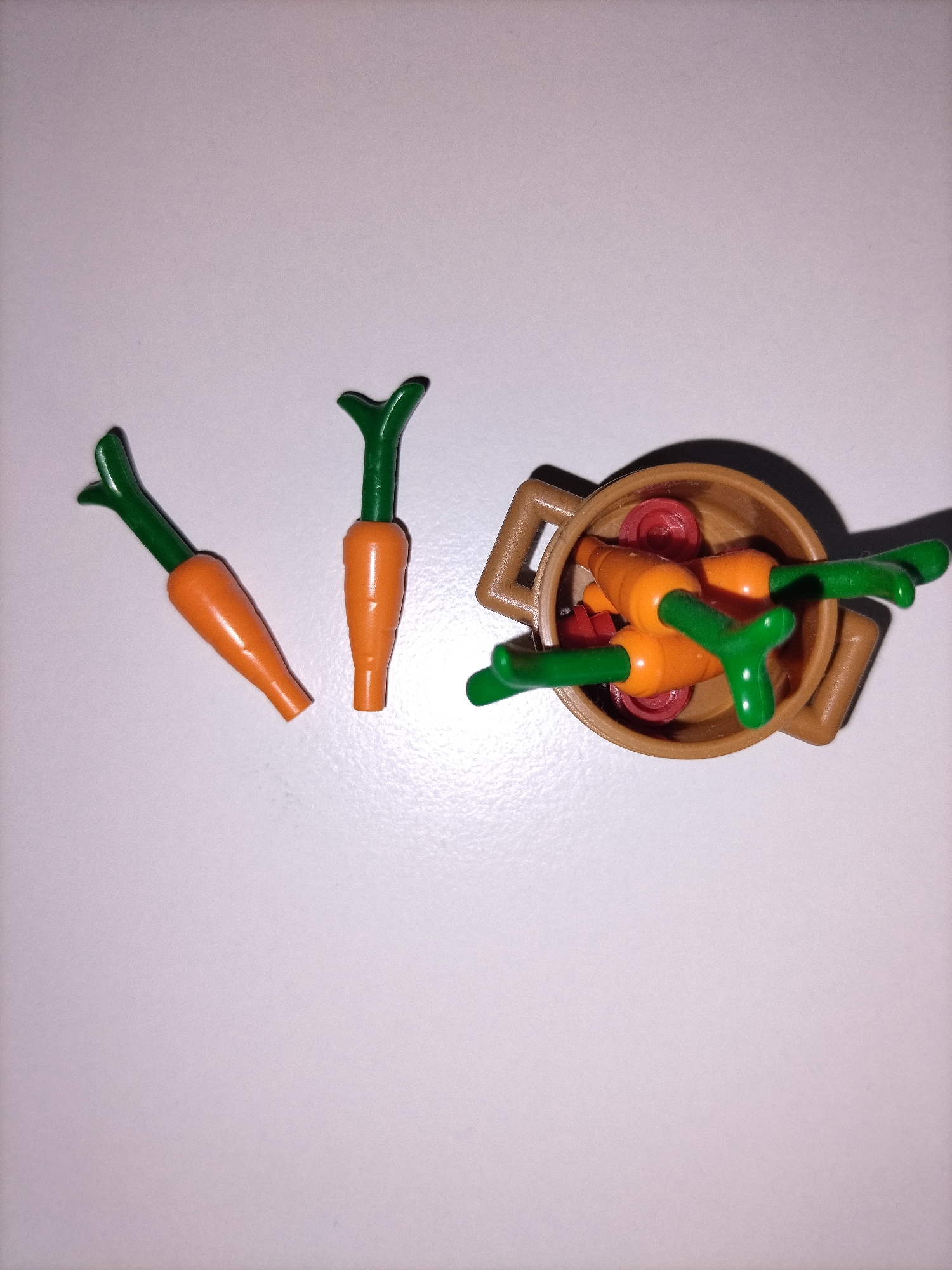 An overhead shot displays a collection of plastic toy carrots and a toy pot on a plain, light grey or off-white surface. To the left, two individual orange toy carrots with green leafy tops are lying flat. To the right, a brown, round toy pot with two handles contains several other toy carrots, also orange with green tops, along with some small, flat, round red toy pieces that resemble radish slices or tomato slices. The carrots inside the pot are oriented in various directions, with their green tops protruding. The pot and the carrots inside cast distinct shadows on the light surface, suggesting an indoor setting with direct lighting. There is no visible text or branding on any of the items. The objects are arranged in a static, still-life composition.