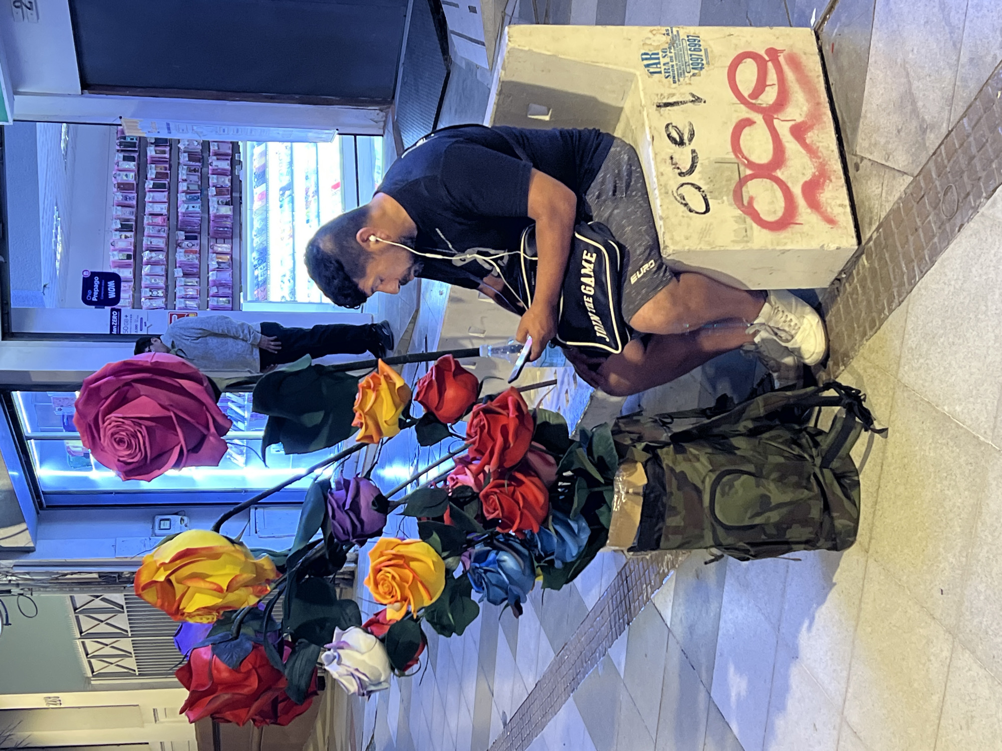 A man kneels on the sidewalk in Arica, Chile, near a concrete block with graffiti and commercial stickers, engrossed in his phone with earbuds in. He rests his elbow on the block, a "JOIN THE GAME" bag at his side, and a camouflage backpack beside him. A bouquet of oversized, colorful roses stands in front of him, contrasting with the store behind him displaying phone accessories. The sidewalk is tiled, and the scene is lit with commercial lighting. Another person lies prone in front of the store.