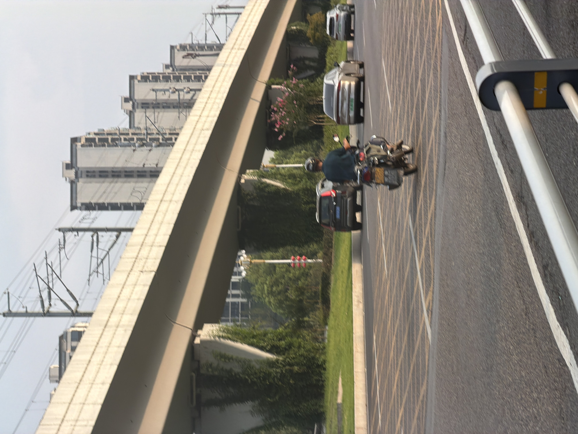 An overpass for an elevated highway or railway runs diagonally across the frame from the upper left to the lower right. Beneath the overpass, on a paved road, a person is riding a motorcycle with a passenger behind them, both wearing helmets. To the right of the motorcycle, a silver sedan is driving in the same direction. Further to the right, another silver sedan is visible. In the background, several tall, modern apartment buildings are lined up. The environment appears to be an urban setting in Jiangjin, China, characterized by extensive infrastructure and residential development.