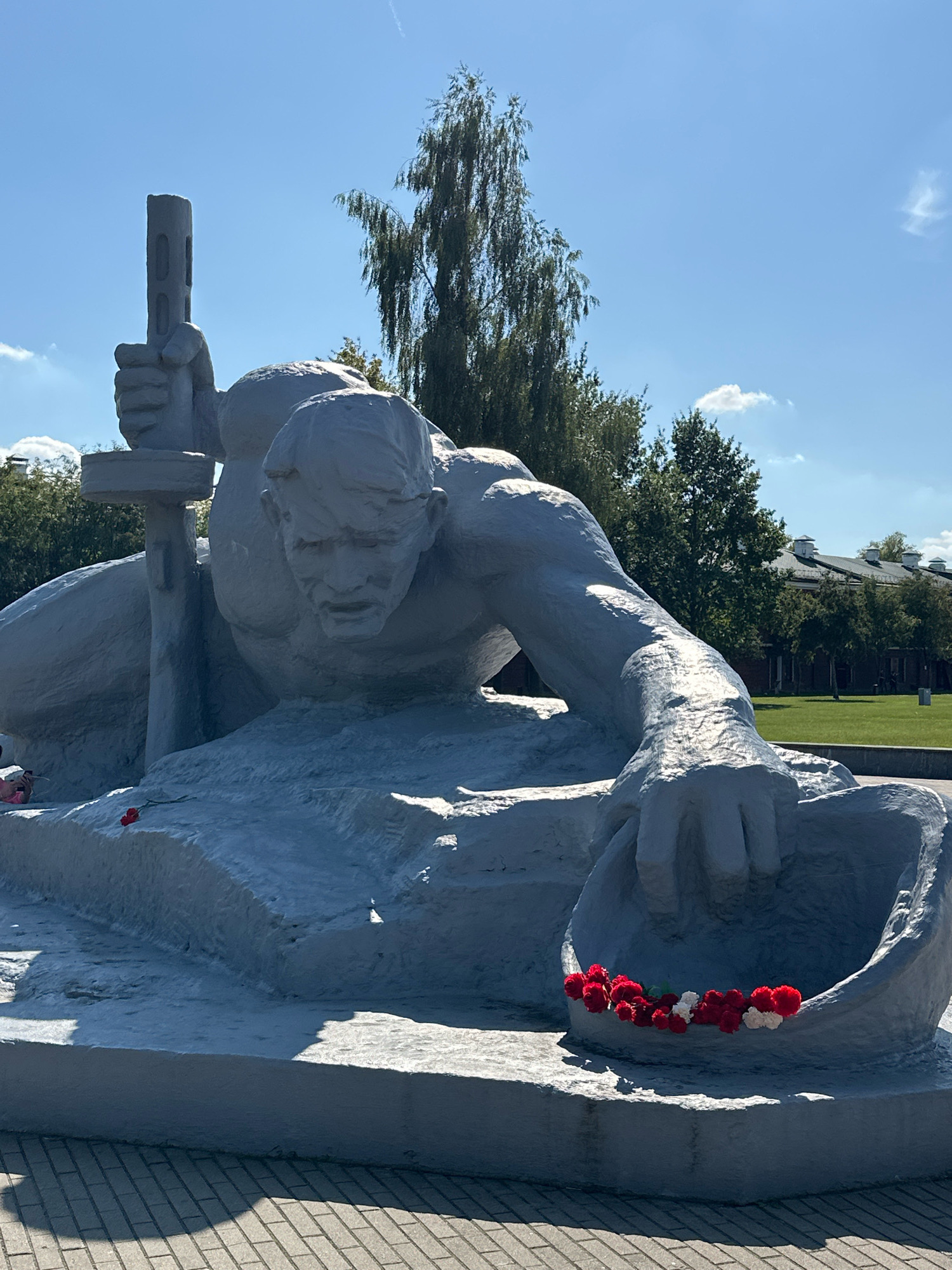 The image shows a statue of a soldier in a prone position, with a helmet cradled in his left hand. The statue is made of concrete and is painted gray. Red and white flowers are arranged in the helmet. The soldier is holding a rifle in his right hand. The statue is located at the Brest Fortress in Brest, Belarus. The fortress is a UNESCO World Heritage Site and a symbol of Soviet resistance during World War II. The sky is clear and blue, with a few white clouds. Trees and buildings can be seen in the background. The ground in front of the statue is paved with bricks.