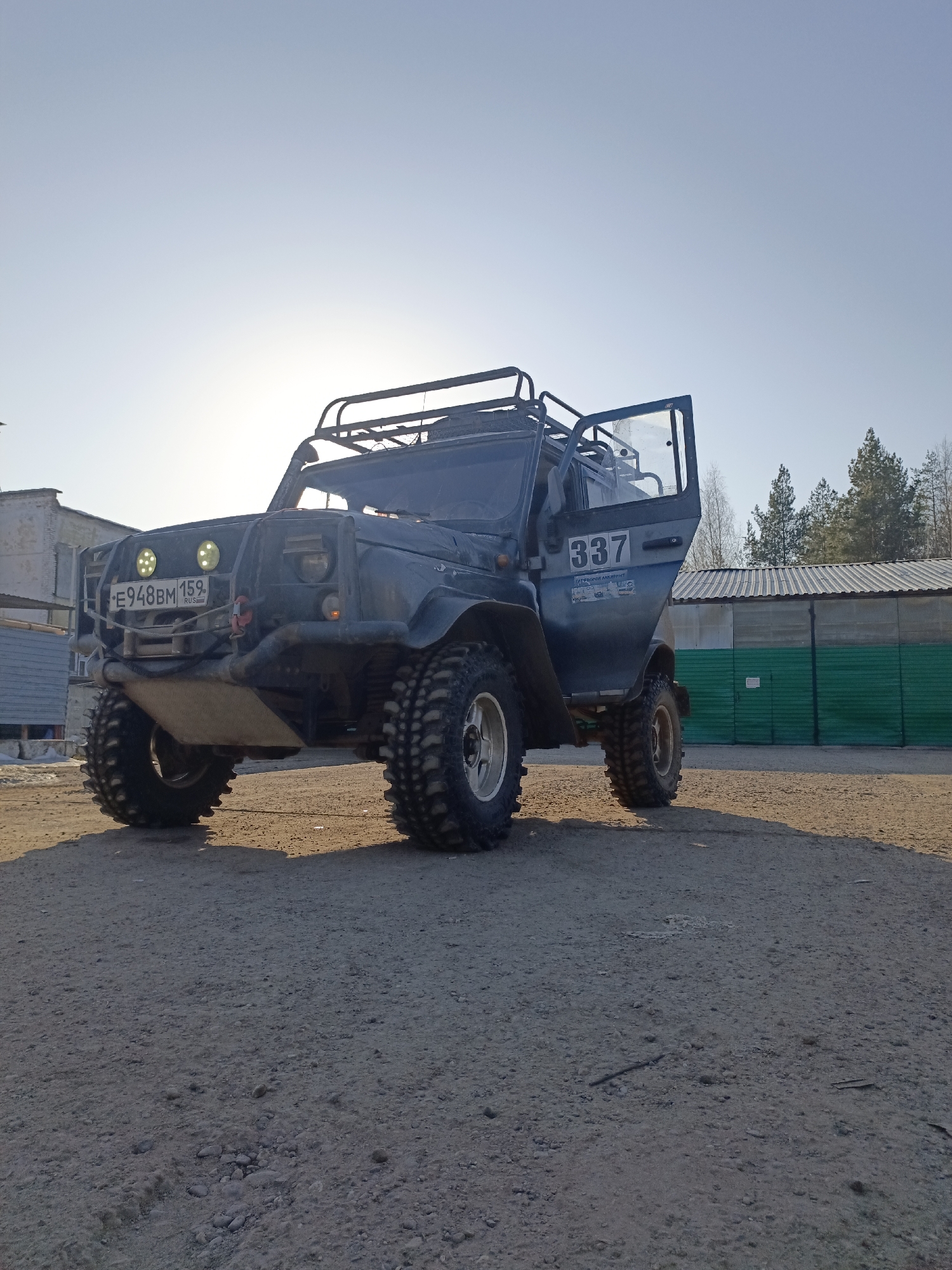 An off-road vehicle is captured in a low-angle shot on an unpaved, dusty ground surface in Noviy, Russia. The vehicle, appearing dark blue or grey, is heavily modified with large, knobby off-road tires on silver rims, a heavy-duty front bumper equipped with a winch, an underbody skid plate, and two circular LED auxiliary lights. A Russian license plate, "E948BM 159 RUS," is affixed to the front. A roof rack is installed on top. The driver's side door is open, revealing a portion of the interior. The number "337" and Cyrillic text, including "СПОРТИВНОЙ АКЦИИ," are visible on the side of the vehicle.
In the background, under a clear, light blue sky with the sun positioned low and to the left, are several structures. To the left of the vehicle, a light-colored building with a corrugated metal roof is partially visible. To the right, a long, low building with a green metal facade and a corrugated metal roof stretches horizontally. Beyond this building, a line of coniferous trees and some bare deciduous trees are present. No people are visible in the scene. Long shadows are cast towards the viewer, indicating a low sun angle.
