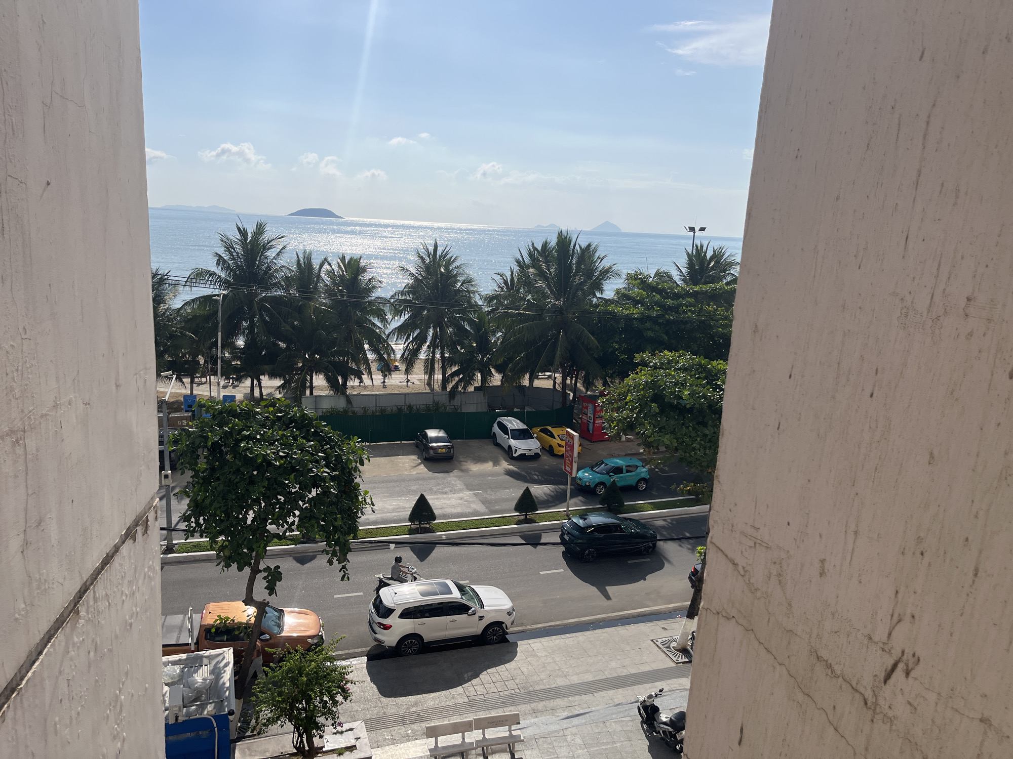 The image shows a street view from an elevated perspective in Bac Nha Trang Ward, Vietnam. Palm trees line the beach across the street. Several parked cars are visible including an orange pickup, a white SUV, a yellow vehicle, and a turquoise hatchback. A dark car is driving on the street. A man on a motorcycle is in the street. A sidewalk with benches and a parked scooter is on the lower right. In the background is an ocean with islands. The image is framed by building structures.