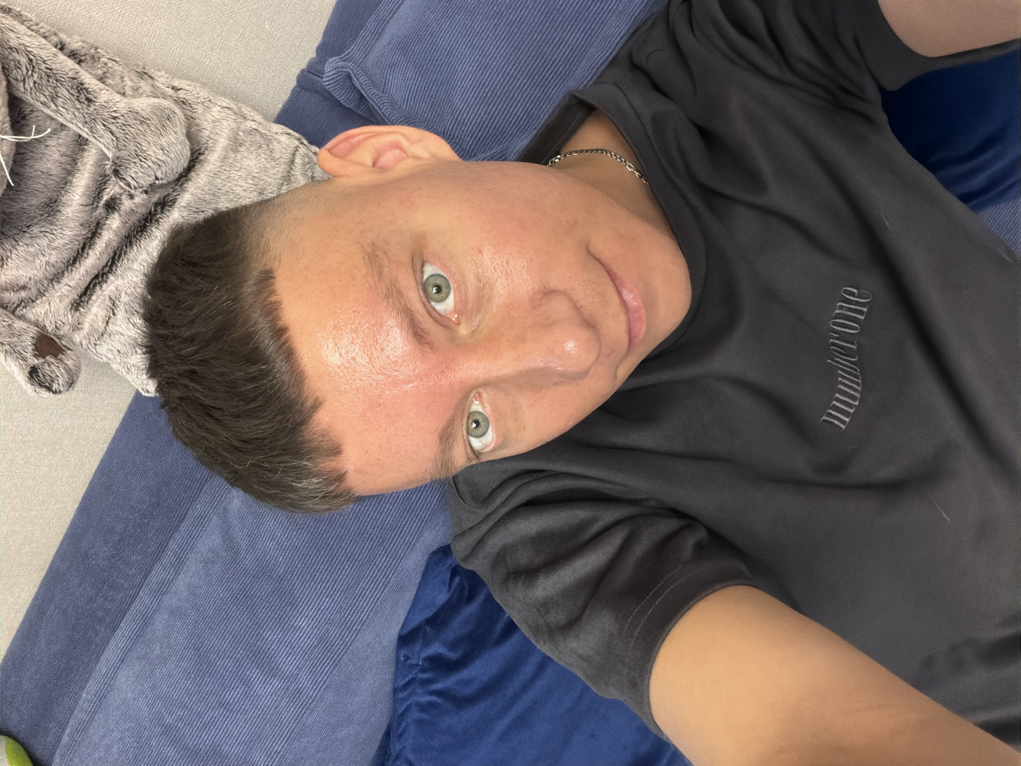 The image shows a close-up, angled selfie of a person lying down. The subject has short, dark hair with a fade haircut and light-colored eyes. They are wearing a dark gray t-shirt with the word "number one" embroidered on the chest. The person is lying on a blue, textured surface, possibly a couch or bed, with a gray, furry blanket to their left. The lighting suggests it's indoors, possibly during the day. The overall mood seems relaxed. No specific details about time of day, weather, or location cues beyond Moscow, Russia are evident.