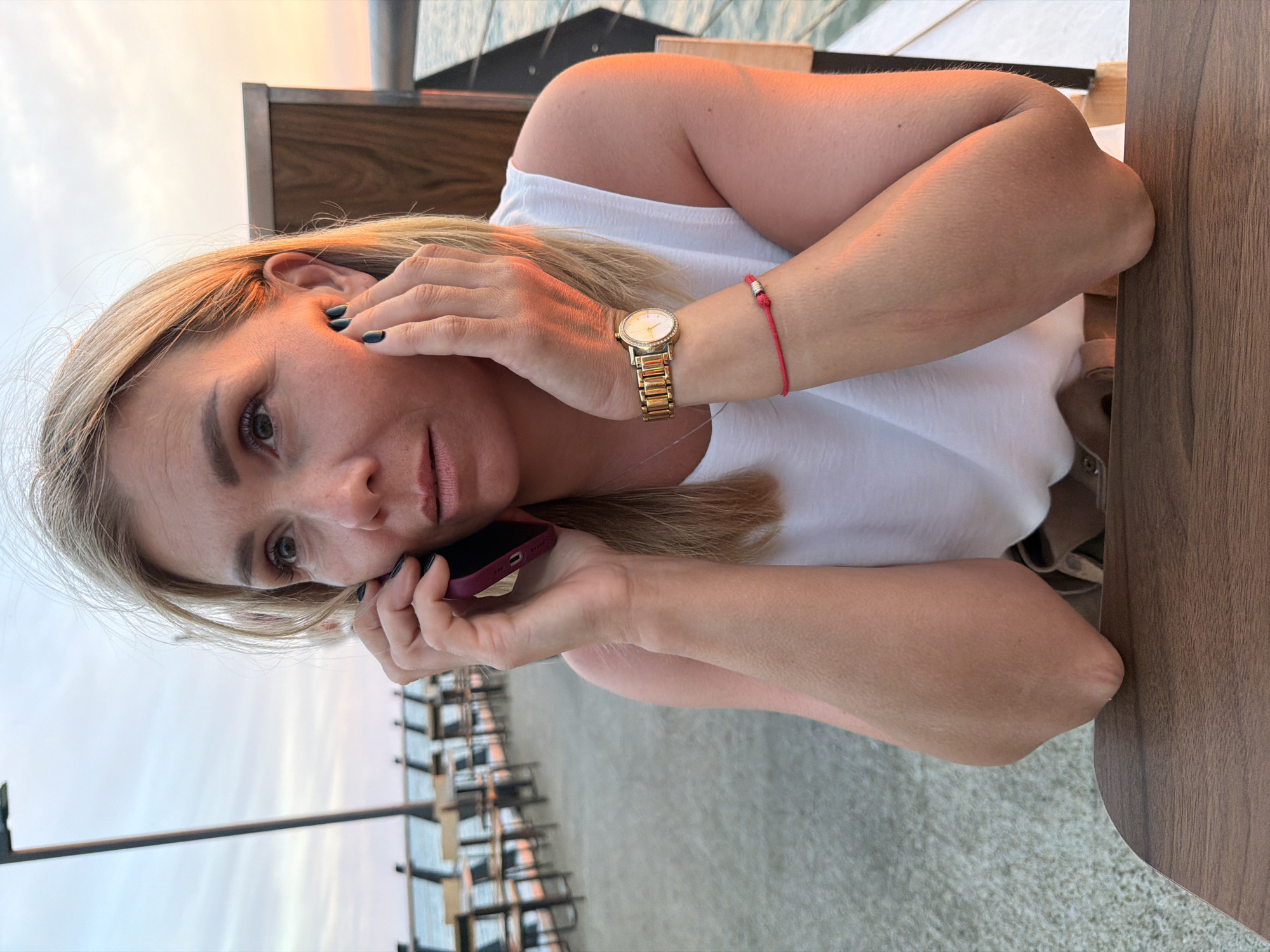 The media file is a portrait-oriented photograph of a woman with blonde hair, lying on her side. She is holding a dark purple mobile phone to her left ear with her right hand, and her left hand is resting near her face, touching her cheek. The woman is wearing a white top and a gold watch with a circular face on her left wrist, along with a thin red bracelet. Her fingernails are painted dark. She is looking towards the camera with a neutral expression.
The setting appears to be outdoors, possibly at a beach or waterfront establishment, given the blurred background showing rows of chairs and what looks like a beach or paved area. The sky in the background is a soft blend of orange, pink, and pale blue, suggesting it is either sunrise or sunset. The lighting on the woman's skin and hair indicates warm, natural light. The background also shows some wooden structures, suggesting she is at a table or lounge area.
The overall mood is relaxed and casual, with the woman seemingly engaged in a phone conversation while enjoying the evening or morning light. There is no visible text in the media file. The location context is Sochi, Russia.