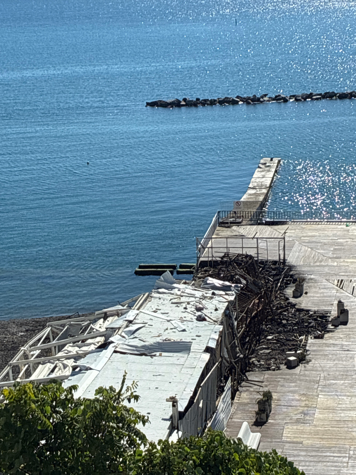 The image shows a partially destroyed wooden structure on a pier by the sea. The structure appears to have been damaged by fire, with debris and burnt materials scattered across the wooden deck. The sea is a vibrant blue with sunlight glinting off the surface. In the distance, a line of rocks forms a breakwater, and a concrete pier extends into the water. The foreground features lush green foliage, suggesting the location is coastal. This scene is located in Sochi, Russia, at the site of the former "Sea Breeze" club, which was destroyed in a fire. There are no people visible in the image.