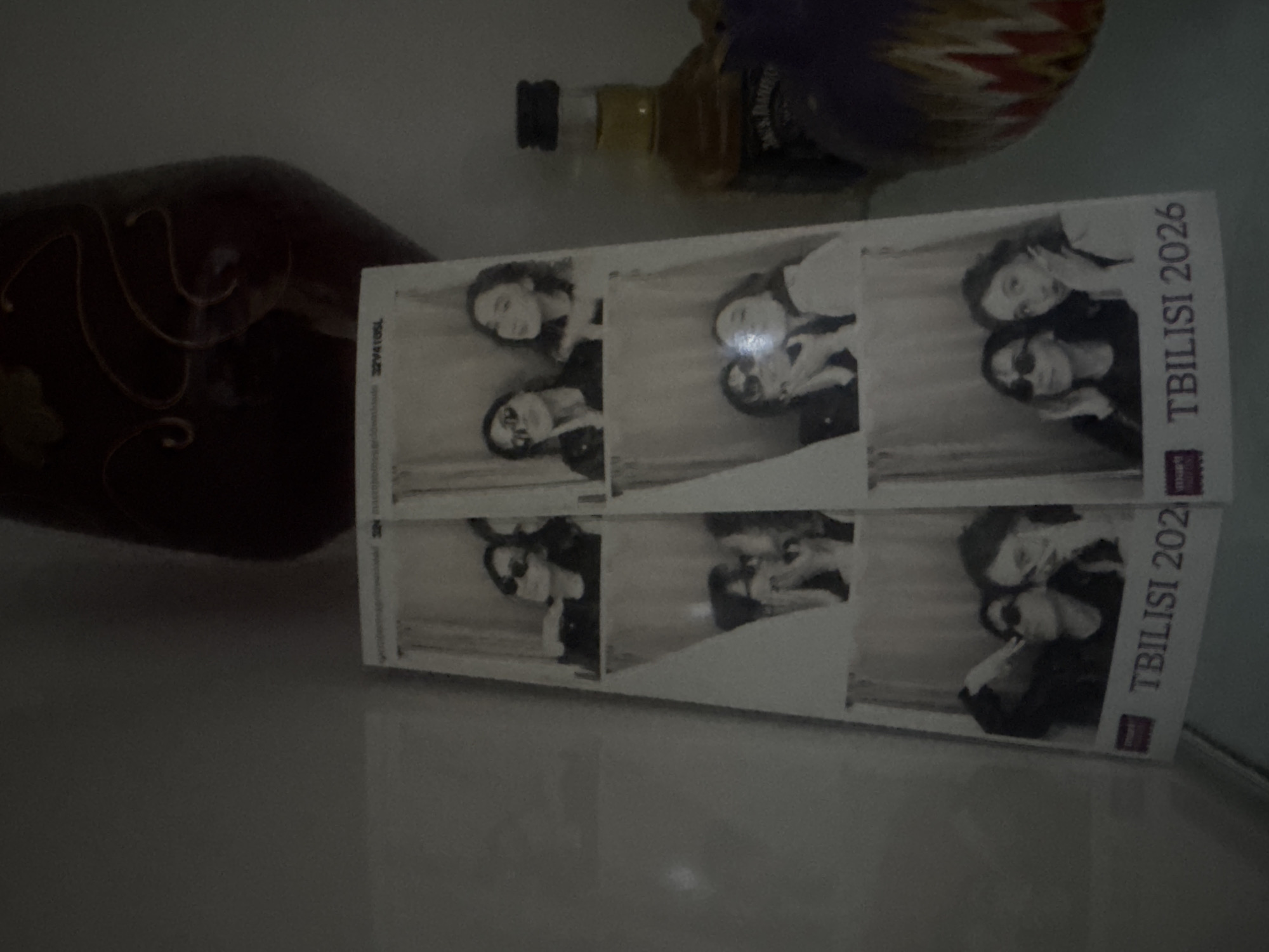 The image shows a dimly lit scene on a reflective surface, possibly a countertop or table. There are three bottles present. One is a brown glass bottle with ornate, embossed designs. Another is a bottle of Jack Daniel's whiskey, and the last has a purple wrapping. In the foreground lies a photo strip, printed in black and white, showing two women posing for a series of photographs. The photo strip is labeled "TBILISI 2026", indicating the location is Tbilisi, Georgia, and the year either of the trip or when the images were taken.
