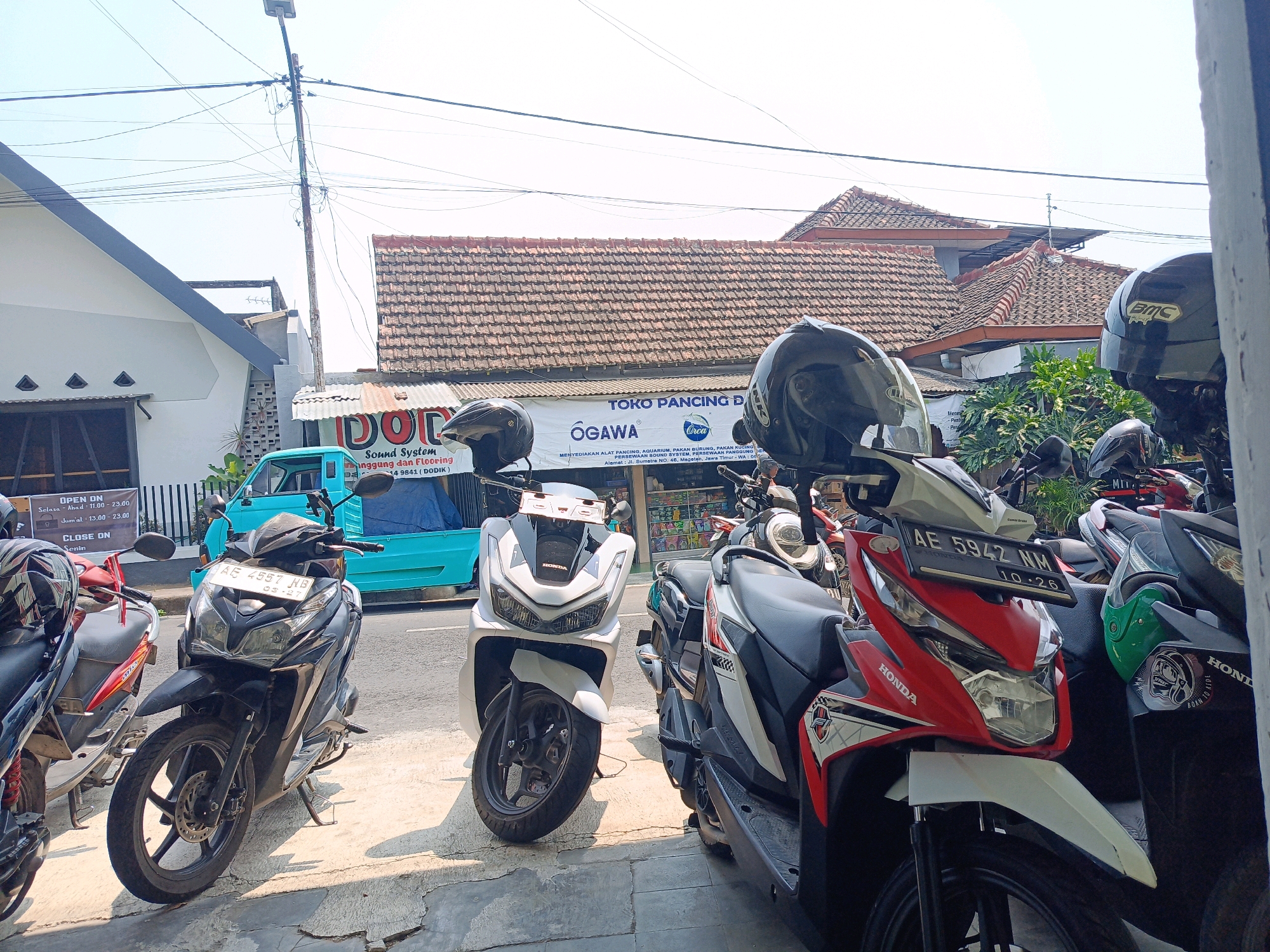 In Kebonagung, Indonesia, a sunlit street scene is captured, featuring a row of parked scooters and motorcycles on a sidewalk. The foreground is dominated by several two-wheeled vehicles, with a black scooter prominently displayed, its license plate reading "AE 4557 NB." To its right, a red and white Honda scooter with the license plate "AE 5942 NM" is parked. Other scooters and motorcycles, some with helmets, fill the space. Behind the parked vehicles, a turquoise utility van is visible, along with shopfronts displaying various goods. A banner with the text "TOKO PANCING D" and "OGAWA" is hung above the shops, suggesting a fishing tackle store. In the background, residential buildings with tiled roofs and power lines with a street light are present. The environment appears to be a typical urban commercial area.