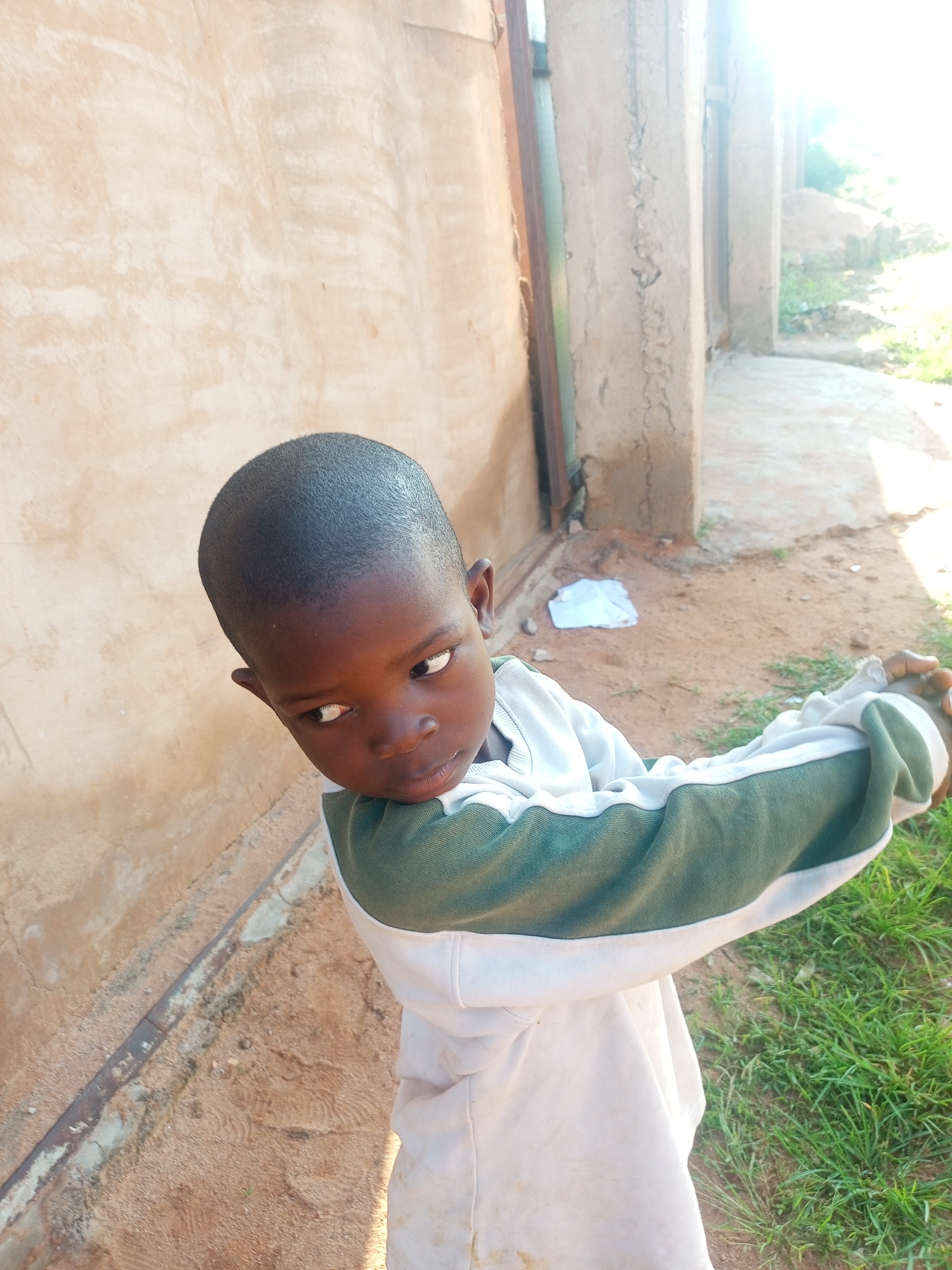 A young Black boy is shown from the shoulders up, looking to his left. He has short, dark hair and is wearing a white sweatshirt with a green stripe across the chest. The background consists of a light brown wall and some green grass. The image appears to be taken outdoors during the day, with bright sunlight. The location is in Tempure, Nigeria.
