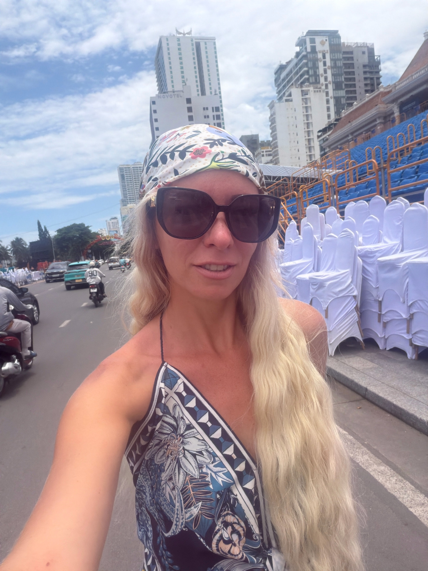 A blonde woman wearing a patterned headscarf and large sunglasses is taking a selfie outdoors. She is wearing a blue and white patterned dress. Behind her, on the right, are rows of white-covered chairs, likely for an event. In the background, there are tall buildings, including the Novotel hotel, and a blue tiered seating area. On the left side of the image, a street scene unfolds with traffic, including motorcycles and cars. The sky is blue with scattered clouds. The location appears to be along a street in Nha Trang, Vietnam, given the architectural style of the buildings and the presence of motorcycles in traffic.