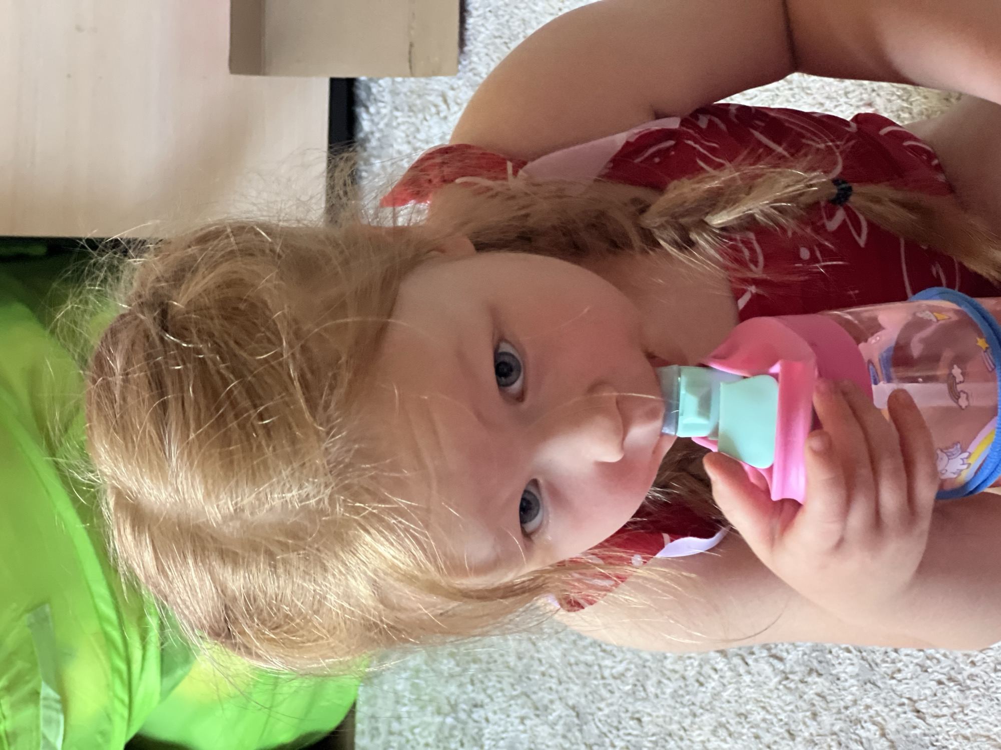 A young girl with blonde hair and blue eyes is drinking from a pink and blue water bottle. She is wearing a red and white patterned dress. The girl is sitting on a carpeted floor in what appears to be a home. In the background, a bright green object and a light-colored surface are visible. The image is taken from a high angle, looking down at the girl. There are no visible indicators of the specific location within the city of Horishni Plavni, Ukraine.
