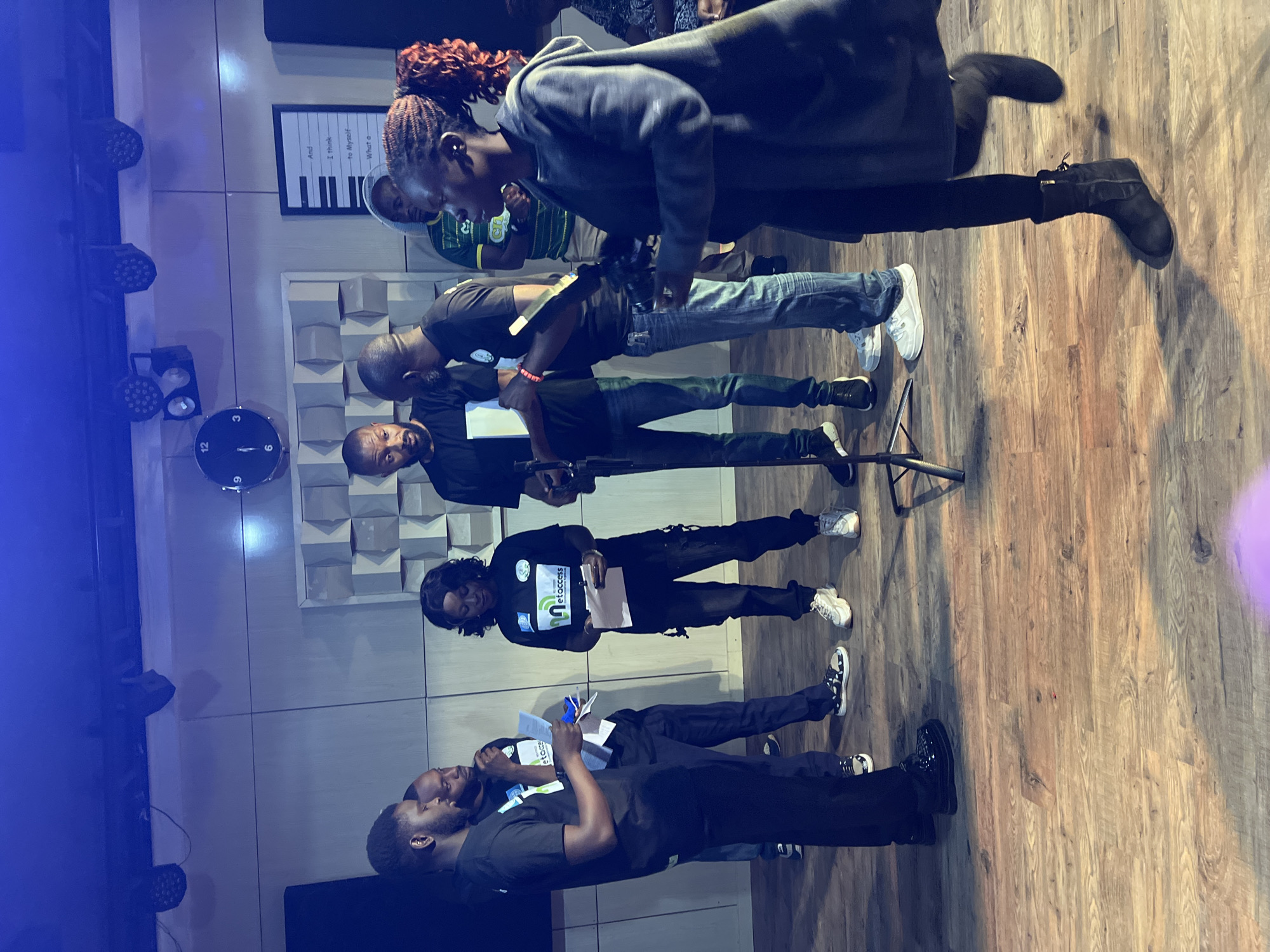 In what appears to be a recording studio or performance space in Jos, Nigeria, a group of seven individuals, six adults and one partially visible person, are gathered. The room is illuminated by blue lighting, with several spotlights visible on the ceiling. A large clock on the wall shows the time as approximately 12:05. There is also a framed print on the wall with text that includes "And I Think to Myself - What a".
The individuals are dressed in a mix of casual and branded attire, with several wearing black t-shirts featuring a logo that appears to say "metaccess". One woman in the foreground is wearing a grey hoodie and black leggings. A man in the center is holding a piece of paper and speaking to the group. Several others are holding papers, suggesting they may be rehearsing or preparing for an event. The flooring is made of wooden planks. A microphone stand is positioned in the foreground.