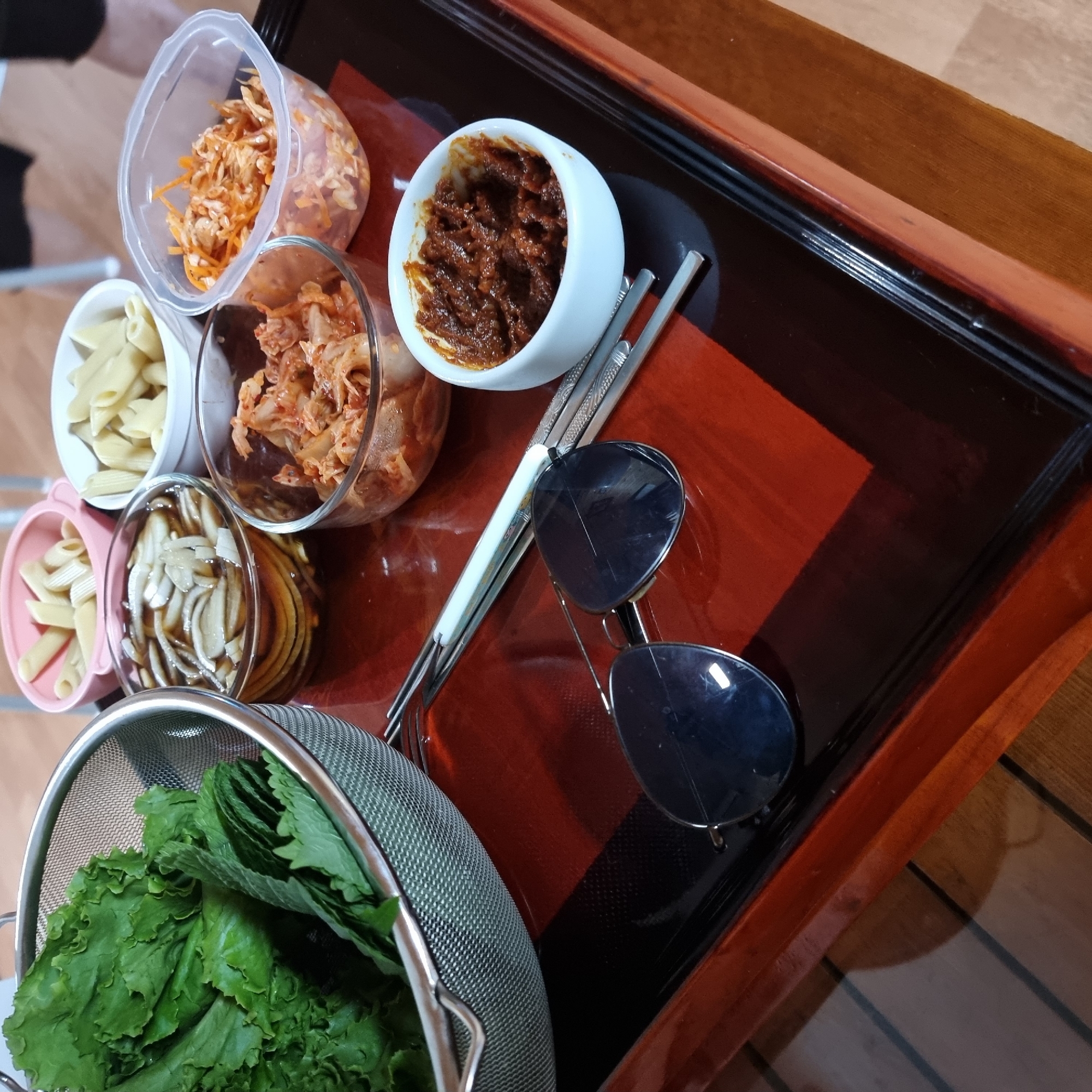 The image displays a selection of food items arranged on a dark, glossy wooden tray with red accents, likely in a residential setting in Dunnae-myeon, South Korea. In a metal colander on the left, fresh green lettuce leaves are visible. To the right, several small bowls contain various food preparations. One pink bowl holds penne pasta. Another white bowl also contains penne pasta. A glass bowl contains sliced mushrooms in a dark liquid. Adjacent to that, a glass bowl holds kimchi. A clear plastic container holds shredded carrots and possibly more kimchi. A small white bowl contains a reddish-brown paste, likely a sauce or condiment. A pair of sunglasses rests on the tray, along with a pair of chopsticks. The lighting suggests an indoor environment, possibly a dining table. No people are present in the image.